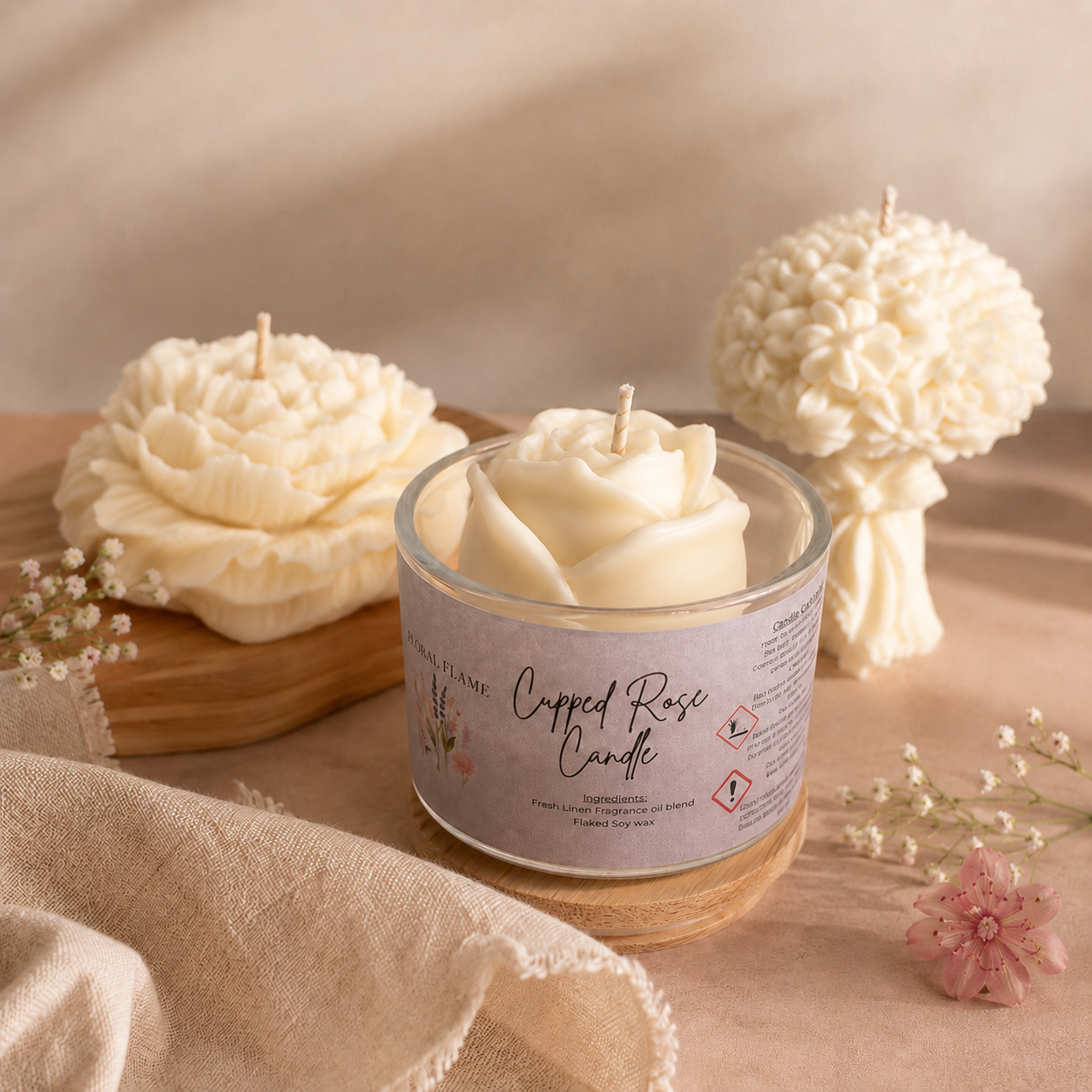 Cream-colored floral-shaped candles arranged on a wooden tray and surface, surrounded by small pink and white flowers, with a beige cloth in the foreground.