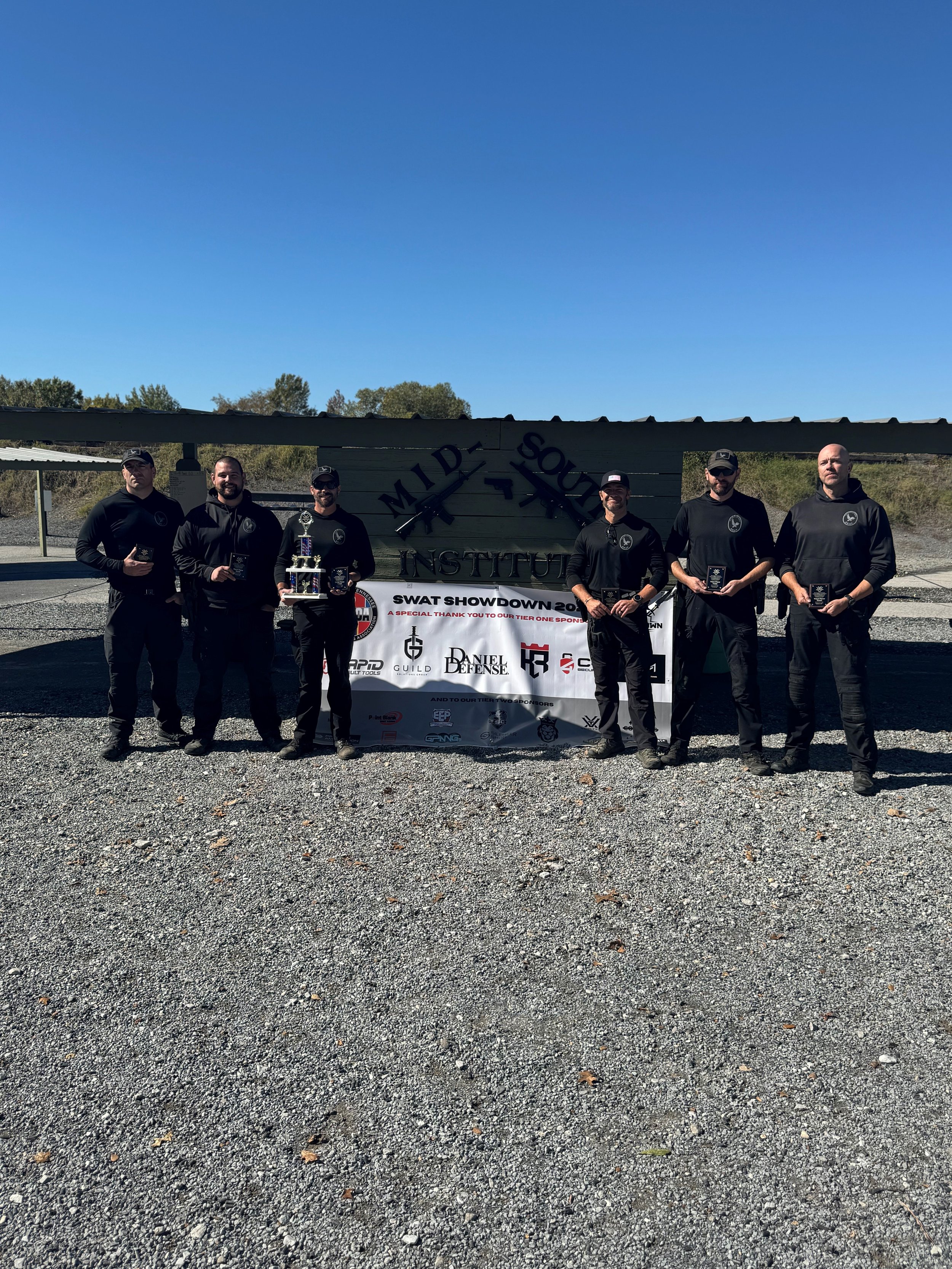 Six men dressed in black, standing outdoors on gravel, holding awards, with a banner and a sign that reads "Mild Forces" and "SWAT Showdown 20" behind them.