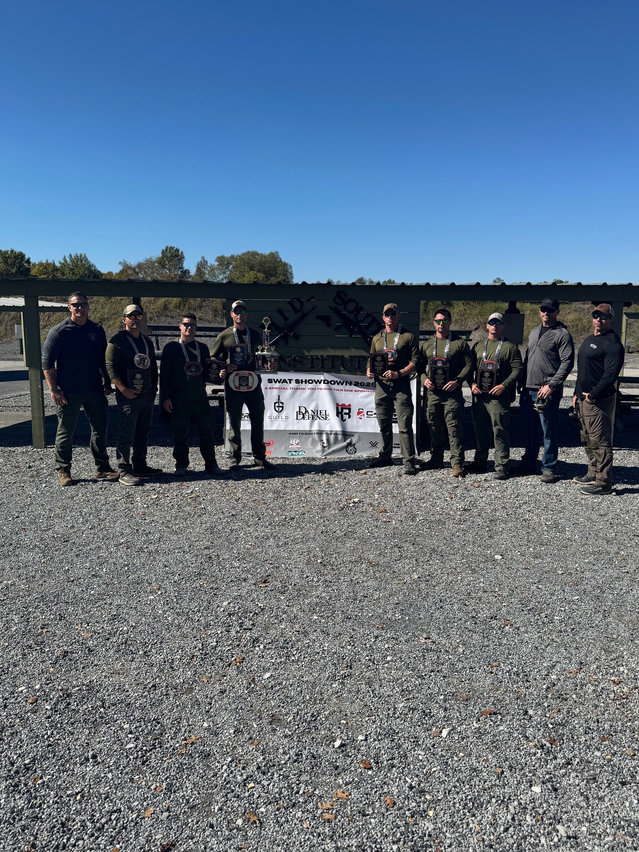 Group of nine men standing on gravel ground in front of a fence, holding plaques, at a SWAT-themed shooting event, with a banner behind them displaying the event name and sponsors, outdoors under a clear blue sky.