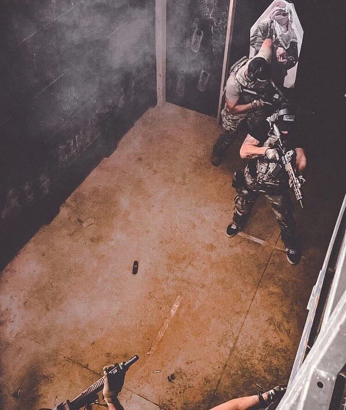 Two children dressed in combat gear and holding toy guns playing in an indoor room with dark chalkboard walls. One is upper right, and the other is lower right, with a black toy gun on the floor.