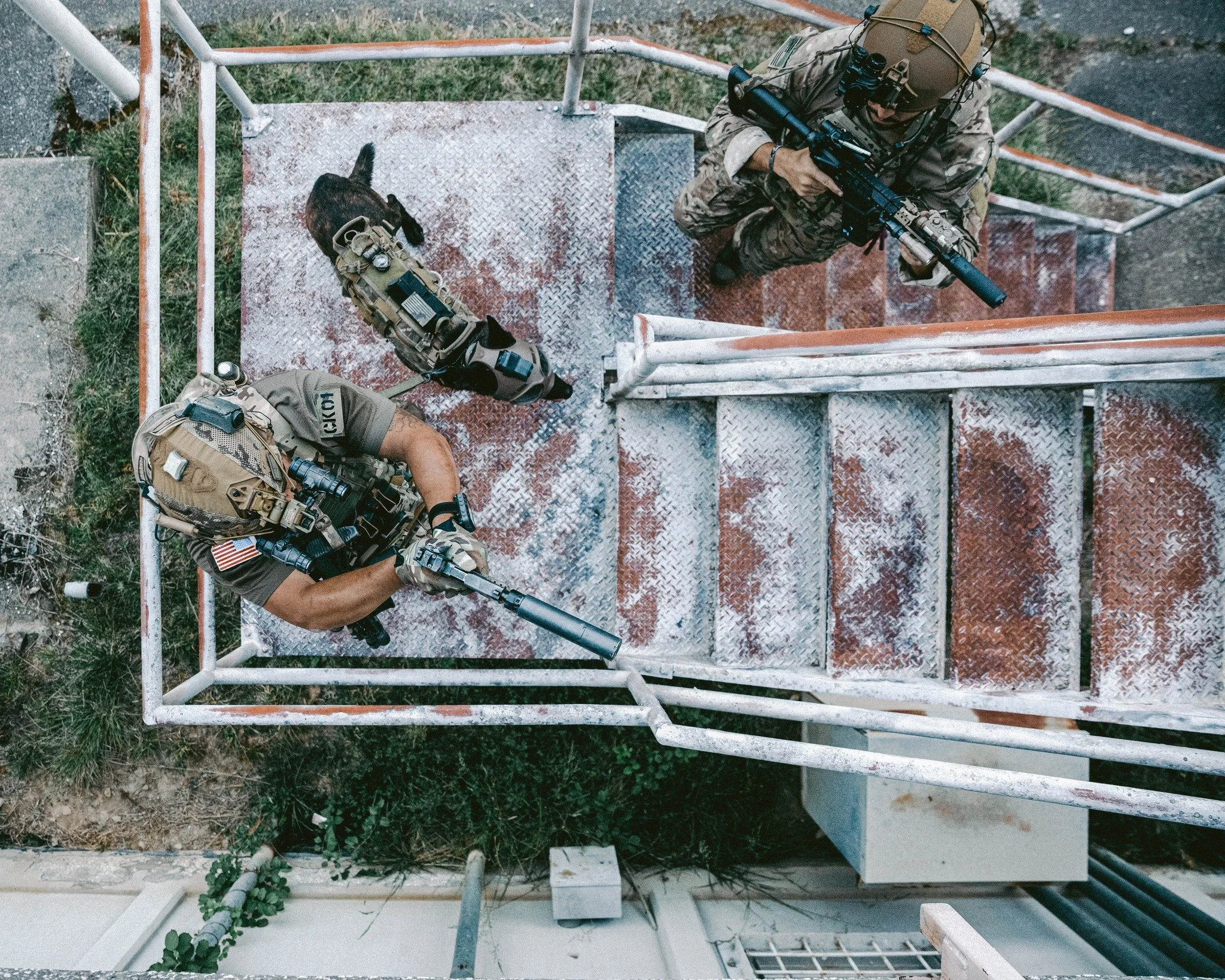 Two armed soldiers in tactical gear on a rusty outdoor metal staircase, looking down from above.