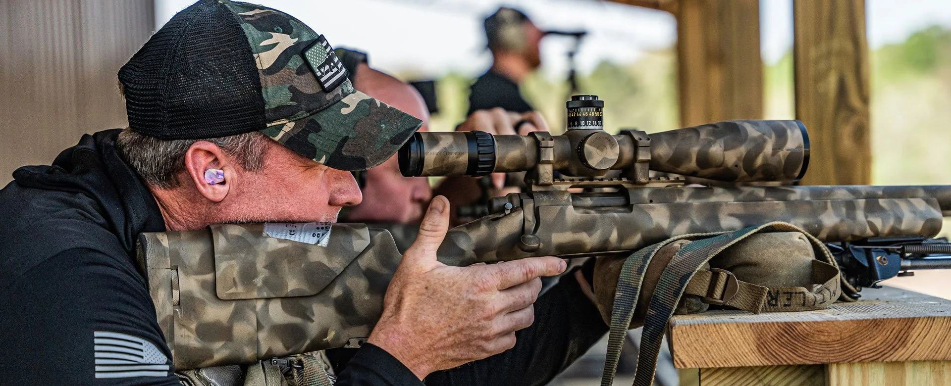 A man aiming a camouflage sniper rifle at a shooting range, wearing a baseball cap and hearing protection, with other shooters in the background.