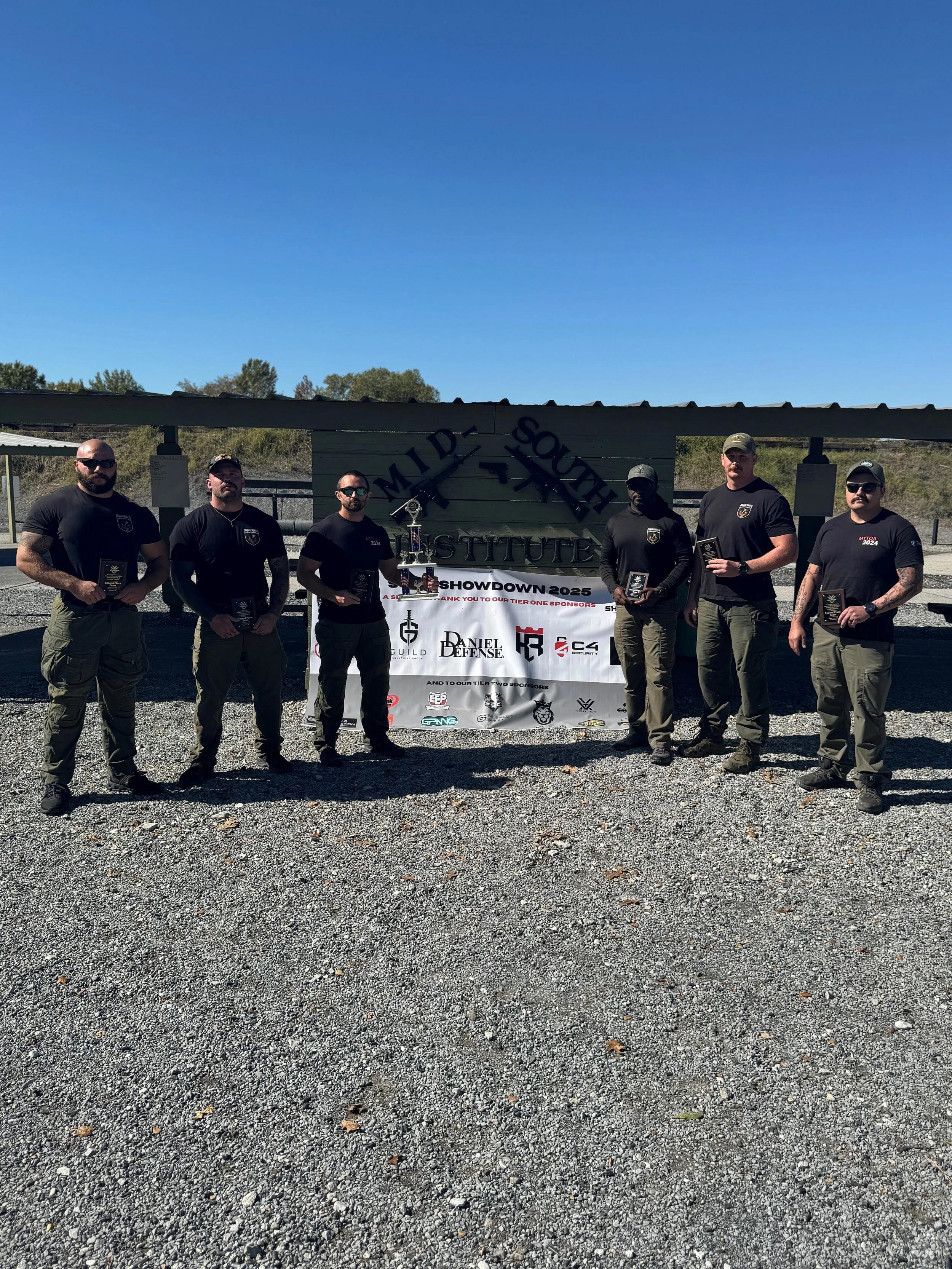 Six men in black shirts and pants standing outdoors in front of a sign that reads 'MIDE SQUARE INSTITUTE' holding plaques, with a banner displaying logos and the words 'SHOWDOWN 2025' below the sign.