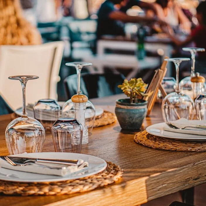 Close-up of a neatly set restaurant table with plates, cutlery, upside-down wine glasses, a small potted plant, and wicker placemats, with diners blurred in the background.