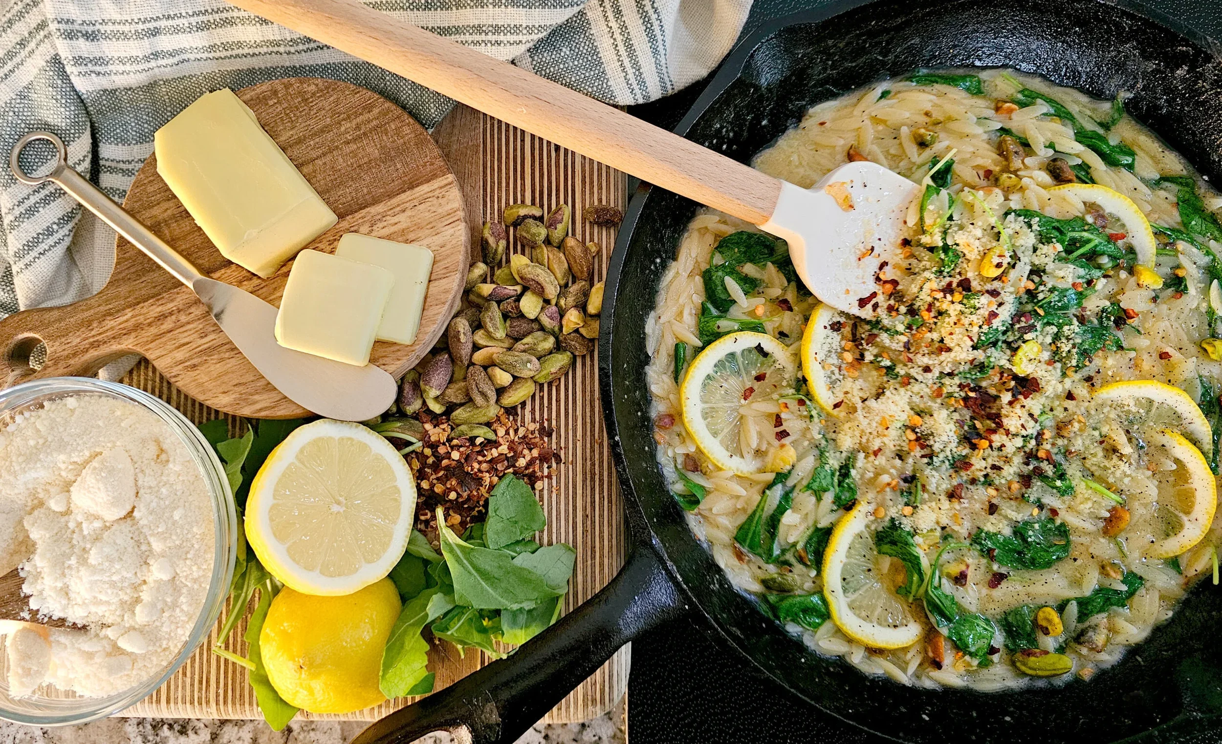 A cast-iron skillet filled with creamy lemon orzo pasta, garnished with spinach, lemon slices, pistachios, red pepper flakes, and parmesan. A spatula rests in the skillet.