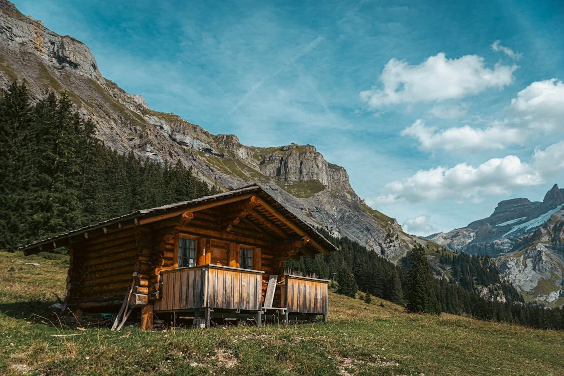 A wooden cabin on a grassy hillside in Durango Colorado with mountains and trees in the background under a blue sky with clouds. Large acreage and ranch appraisals available.