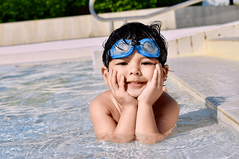 little boy smiling with head in hands wearing goggles and sitting in shallow water