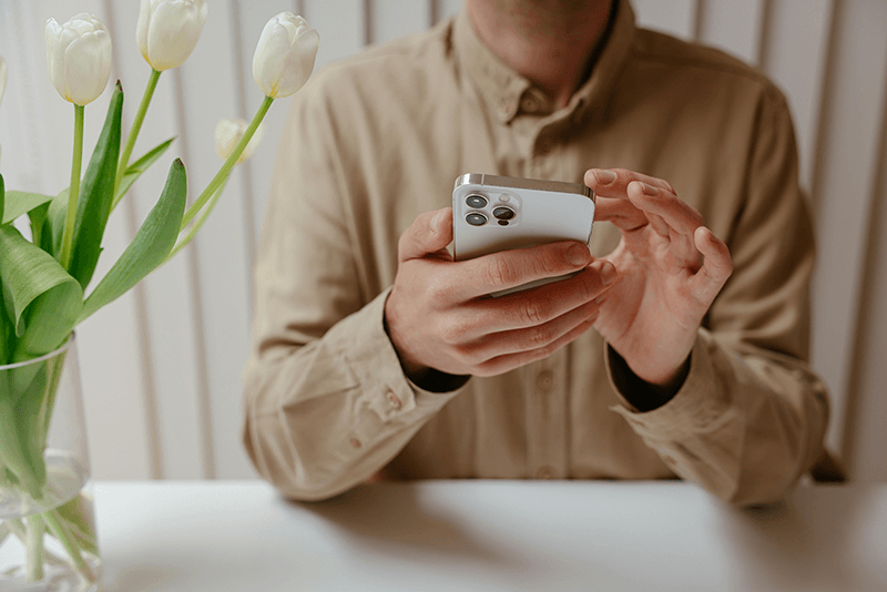 person in tan shirt holing phone with tulips in vase beside them