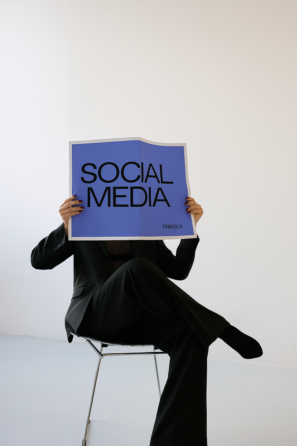 Person dressed in a black suit sitting on a modern chair, holding a large blue sign that reads 'Social Media' in bold letters, with a plain white background.