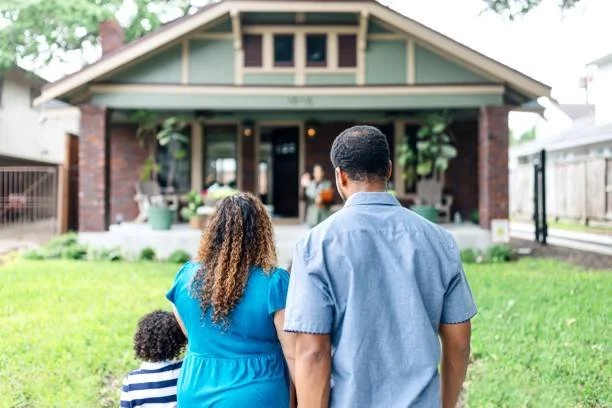 A family of three walking towards a house with a green lawn in front. The family includes an adult woman, an adult man, and a small child, viewed from behind.