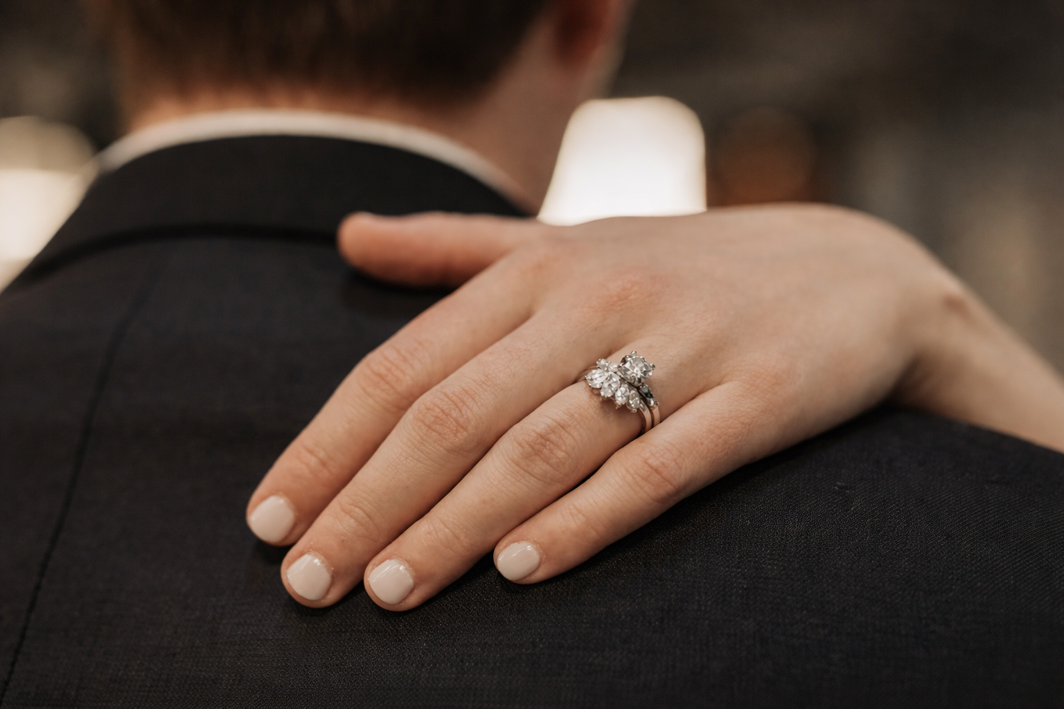 Close-up of a woman's hand with a sparkling diamond ring resting on a man's shoulder, both wearing formal clothing at a romantic occasion.