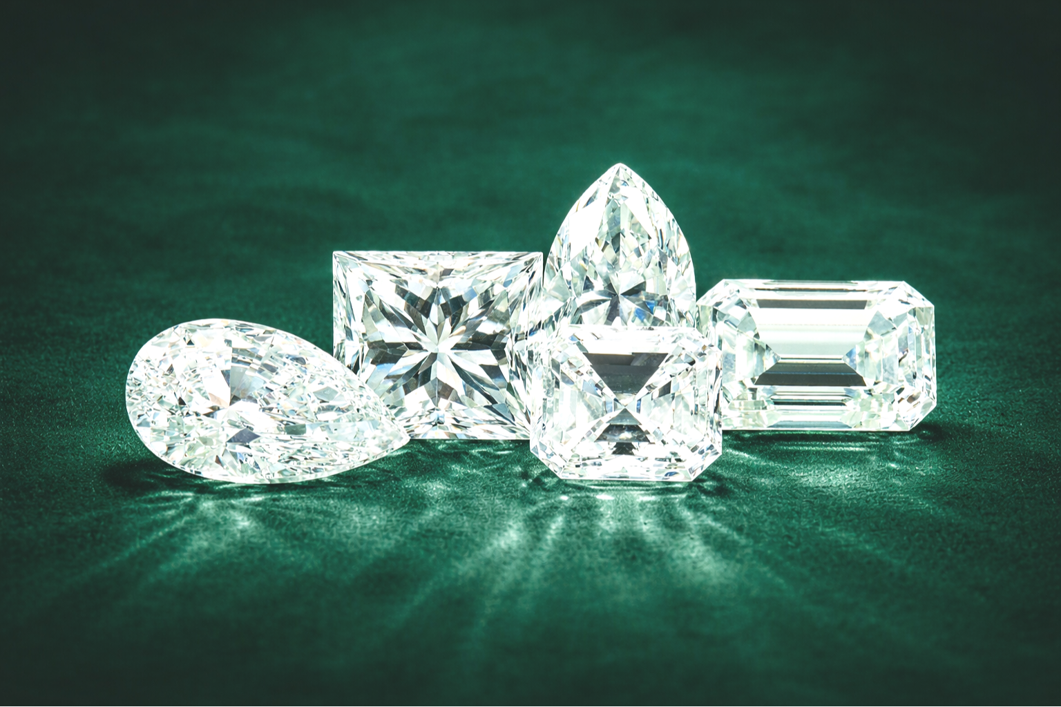 Multiple cut diamonds with various shapes, including oval, heart, square, emerald, and marquise, arranged on a green surface.