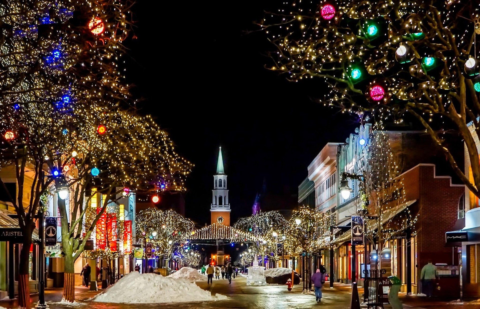 A festive street scene at night decorated with Christmas lights. There are trees wrapped with string lights and colorful ornaments, and snow on the ground. People are walking along the street with storefronts illuminated, leading to a church with a tall steeple in the background.