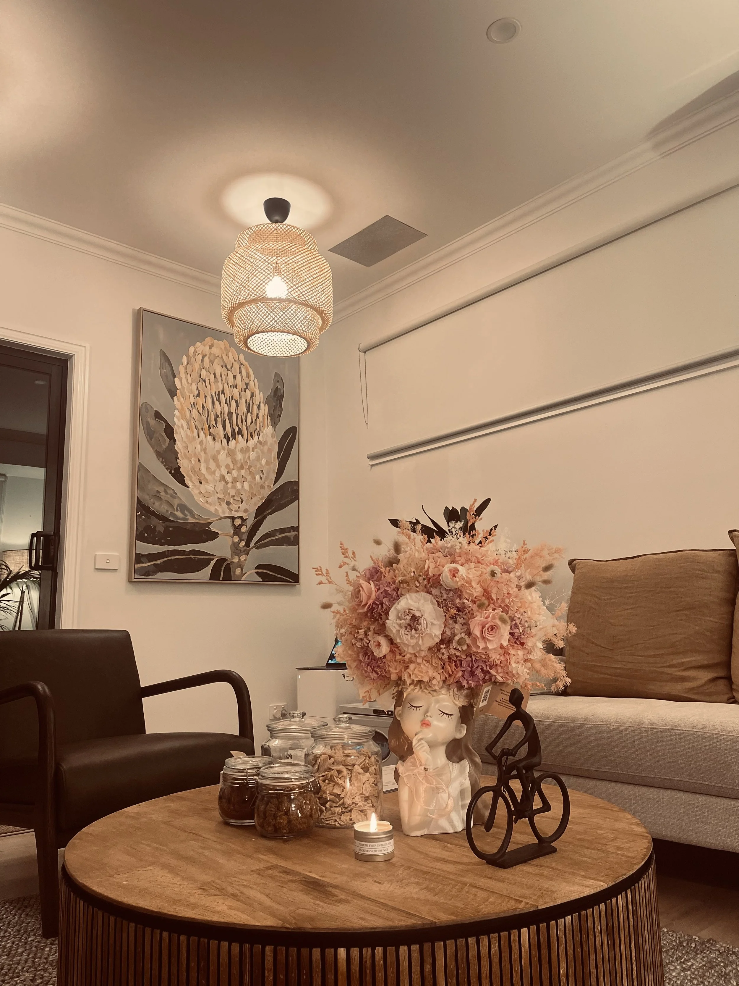 Living room with a round wooden coffee table, a plant-shaped flower arrangement in a white face-shaped vase, jars of snacks, and decorative items. A beige sofa with pillows, a black armchair, wall art of a protea flower, hanging ceiling light, and a window with a roller shade are also visible.