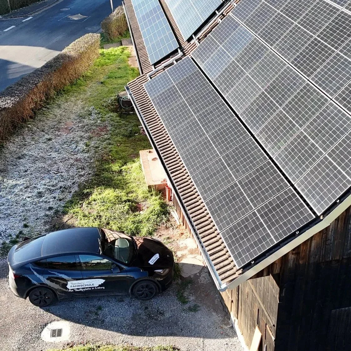 Vogelperspektive auf ein Haus mit Solarzellen auf dem Dach, ein Fahrschul-E-Auto darunter lädt gerade den Sonnenstrom in den Akku, und umgeben von Garten und Zaun.