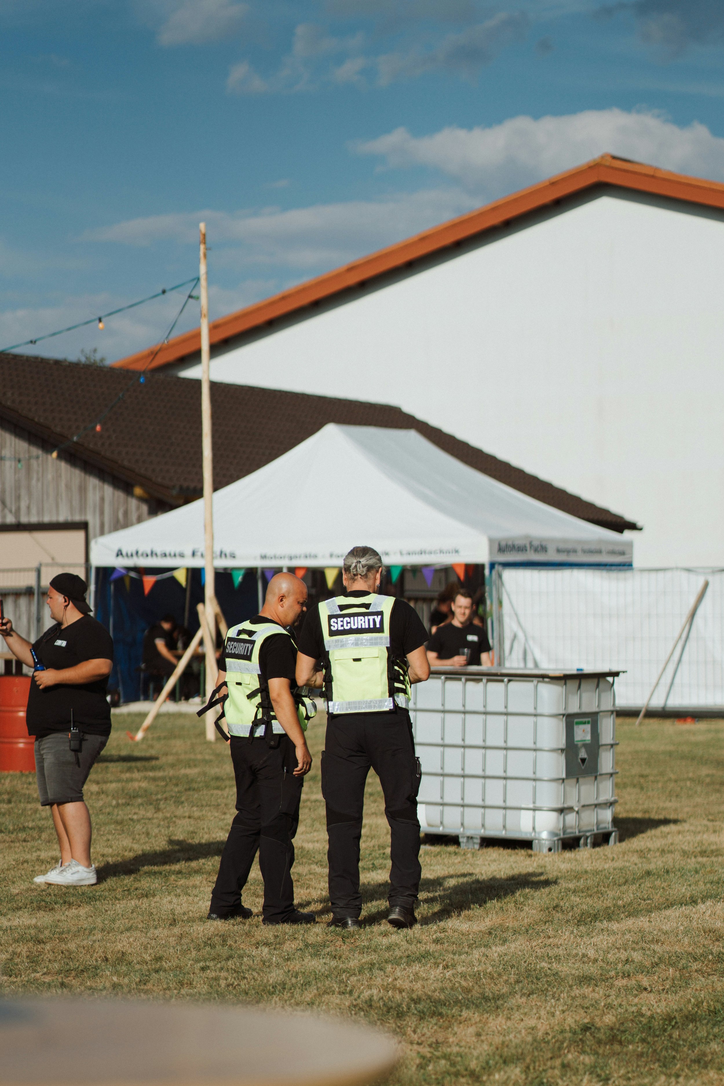 Two security officers with reflective vests standing outdoors near a large trash container, with a white tent and a few people in the background during daytime.