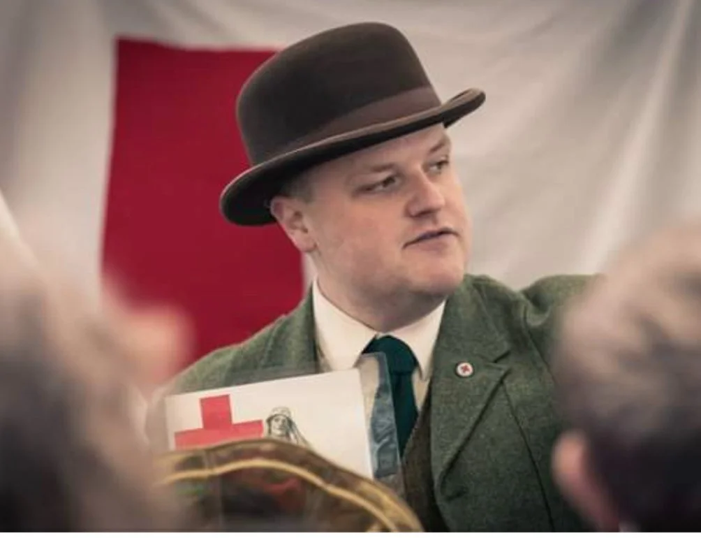 Jesse wearing a vintage green blazer, a tie, and a brown hat with a wide brim, holding a sign with a red cross symbol. He is speaking to a group of people at a WWI history event at a state park in Nashville TN.