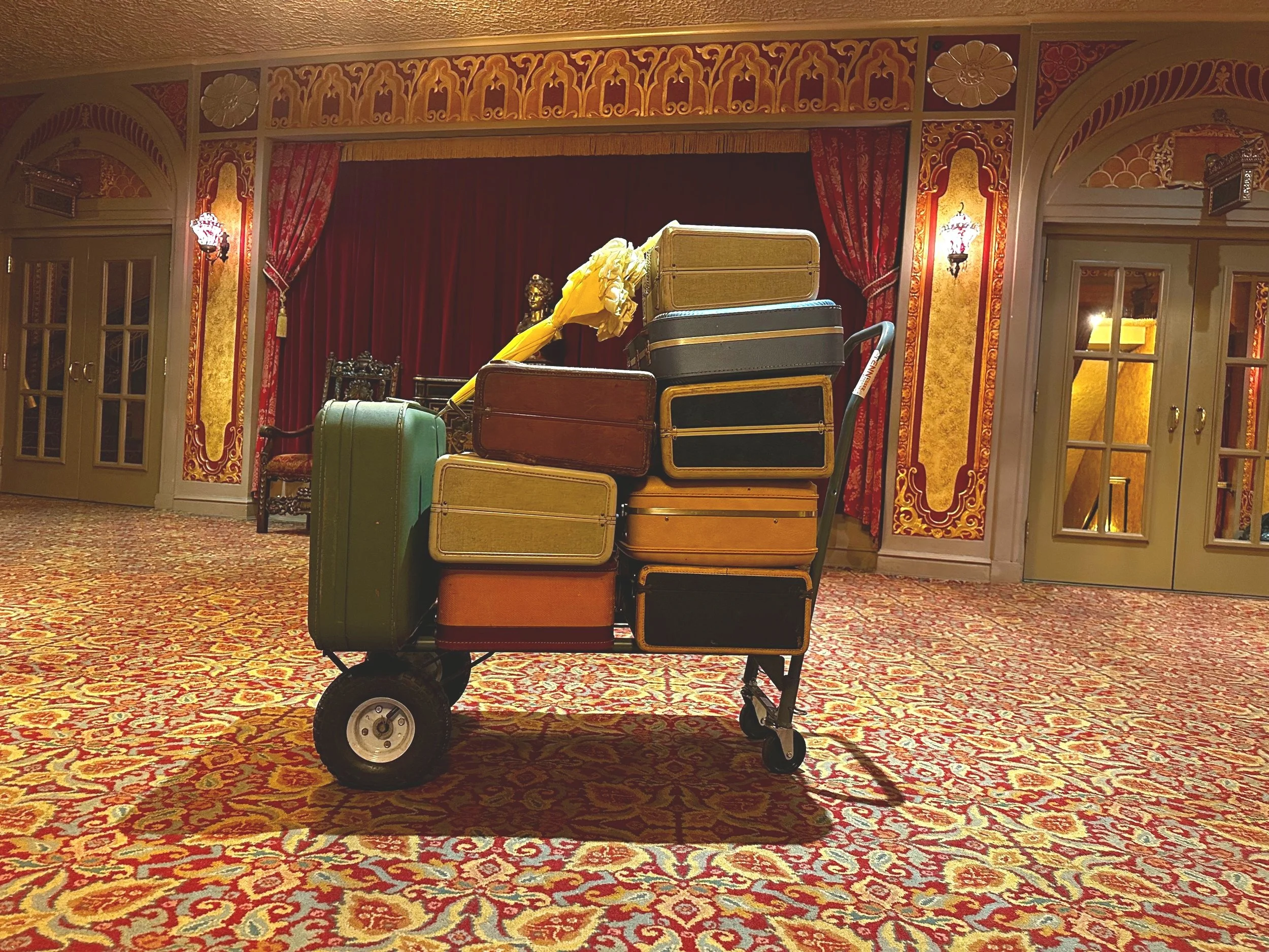 A luggage cart filled with suitcases and an umbrella in the ornate Tennessee Theatre lobby with red and gold decor.