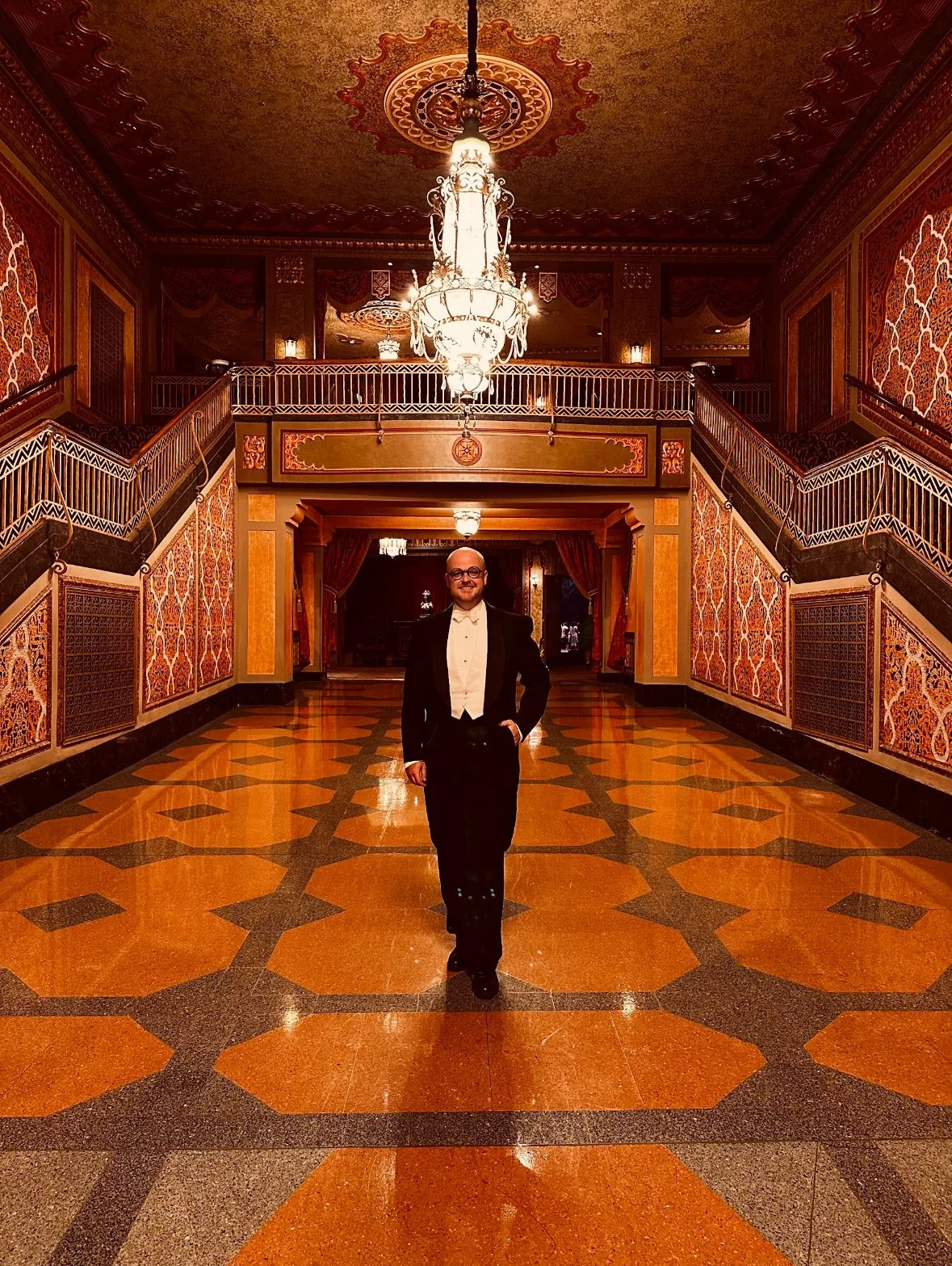 A Jesse in a  1920's tuxedo walking in an ornate, historic lobby with patterned floors, intricate wall and ceiling decorations, and a large chandelier.