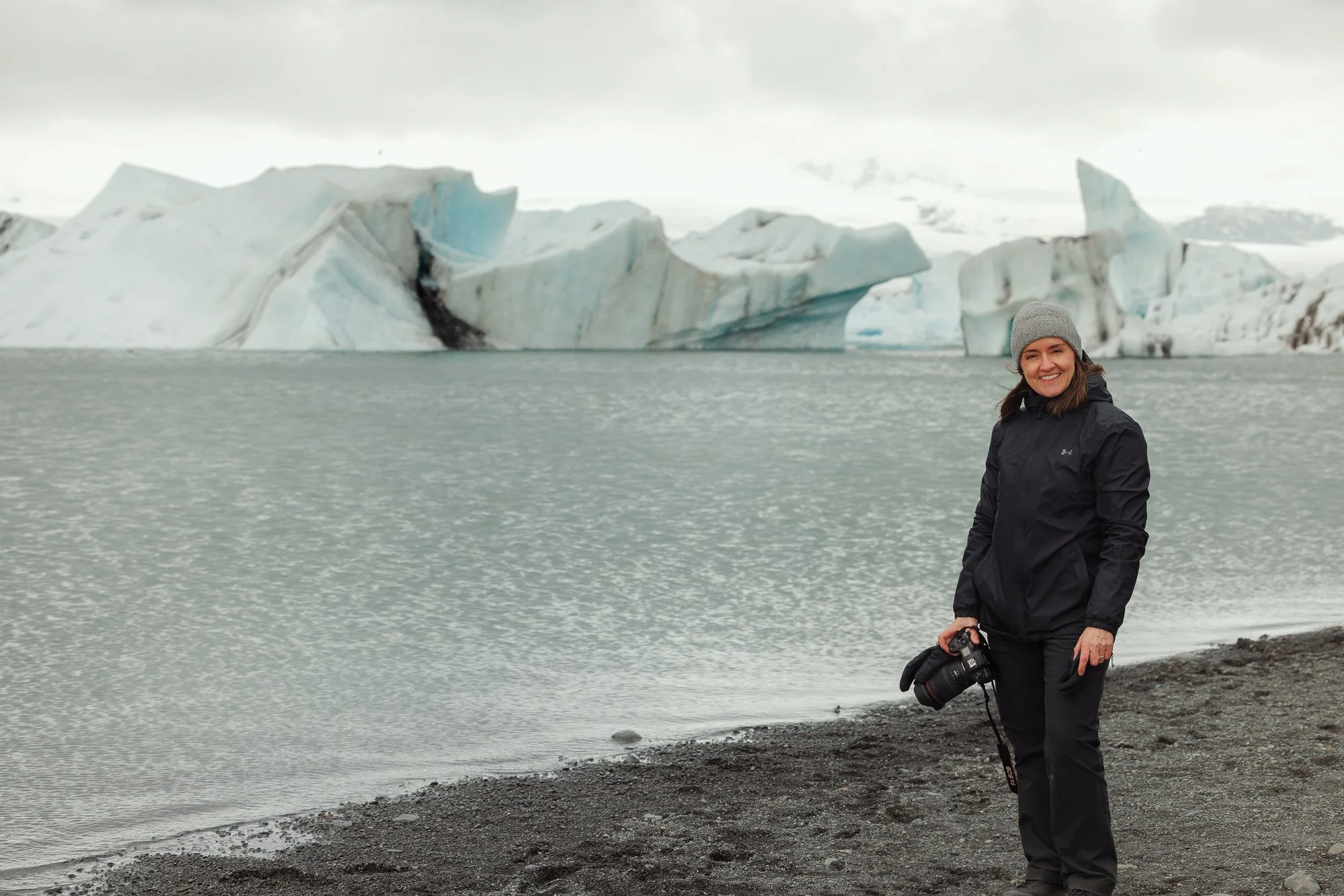 A woman in outdoor winter clothing stands on rocky shoreline beside water, holding a camera, with large icebergs and snow-covered landscape in the background under cloudy sky.