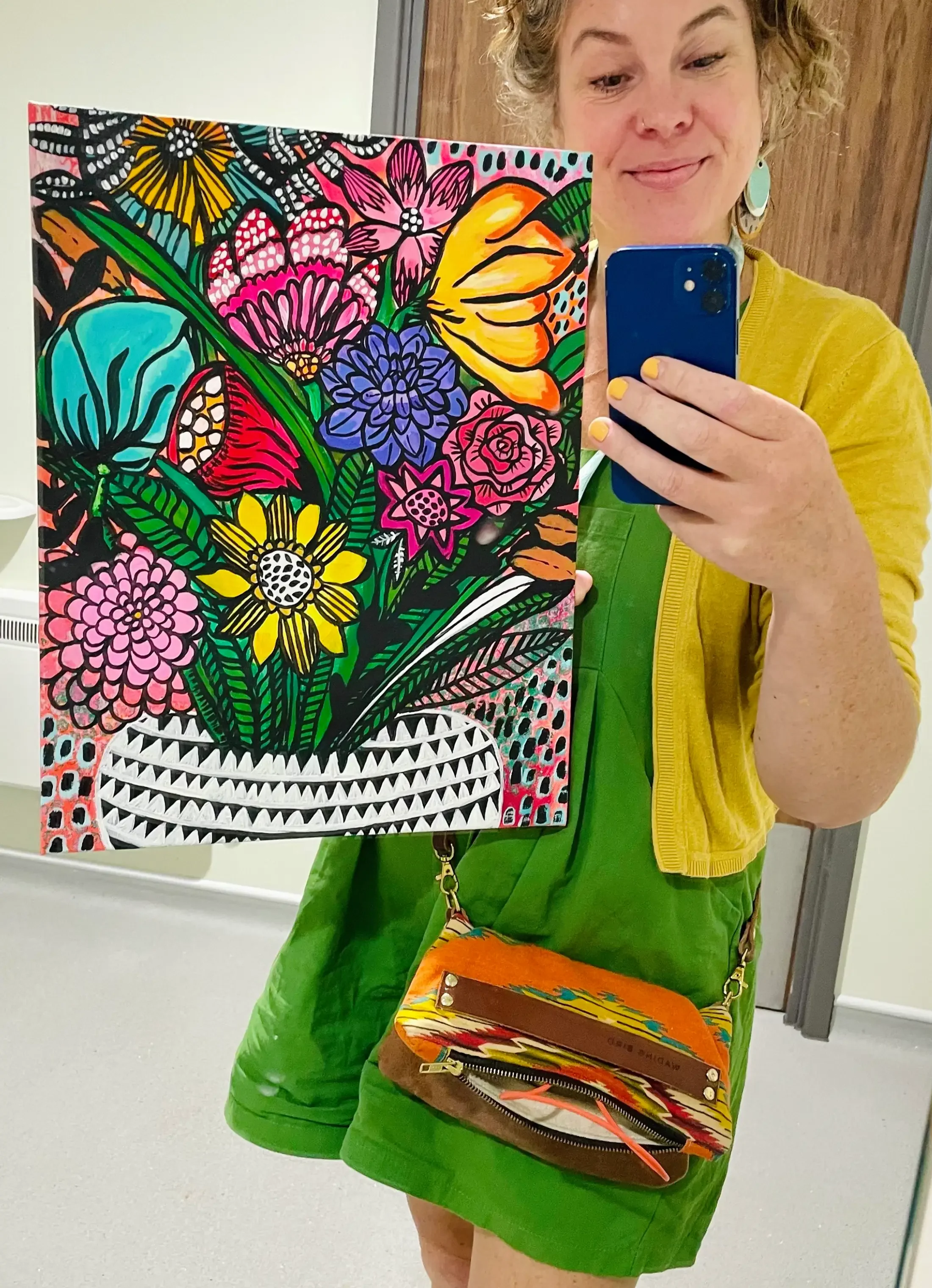 Woman in green dress and yellow jacket holding a colorful floral painting and taking a selfie with her phone.