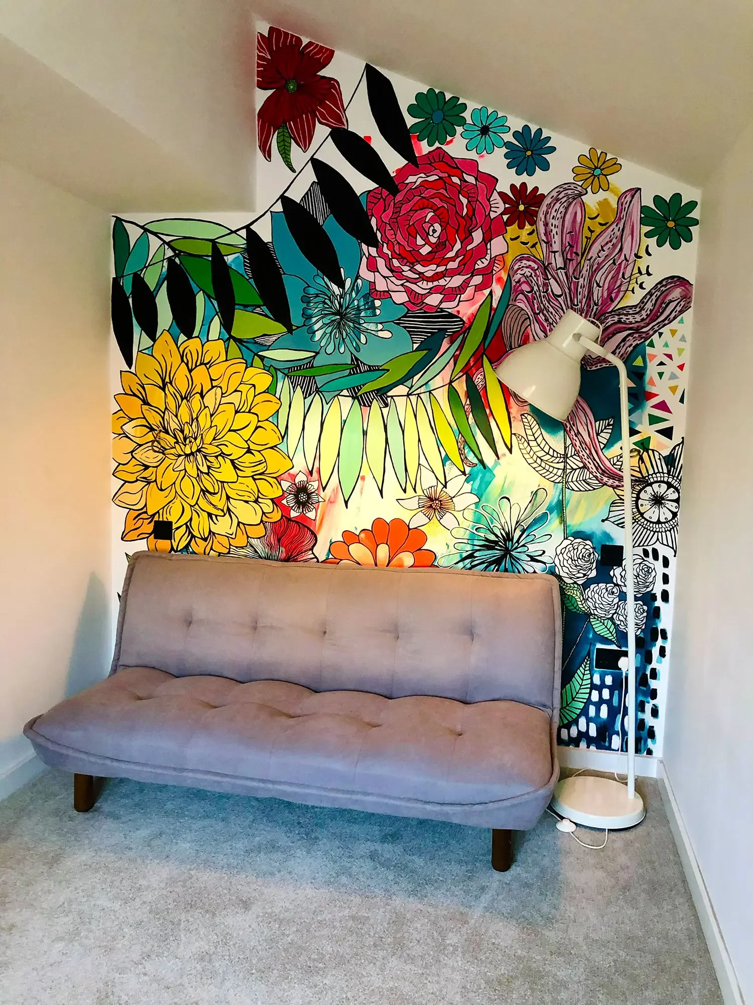 A pink sofa in front of a colorful floral mural wall with a white floor lamp beside it.