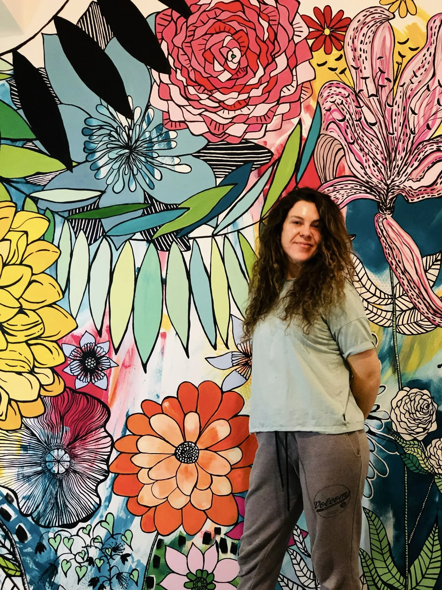 A woman with curly hair standing in front of a colorful, large floral mural featuring various stylized flowers and leaves.