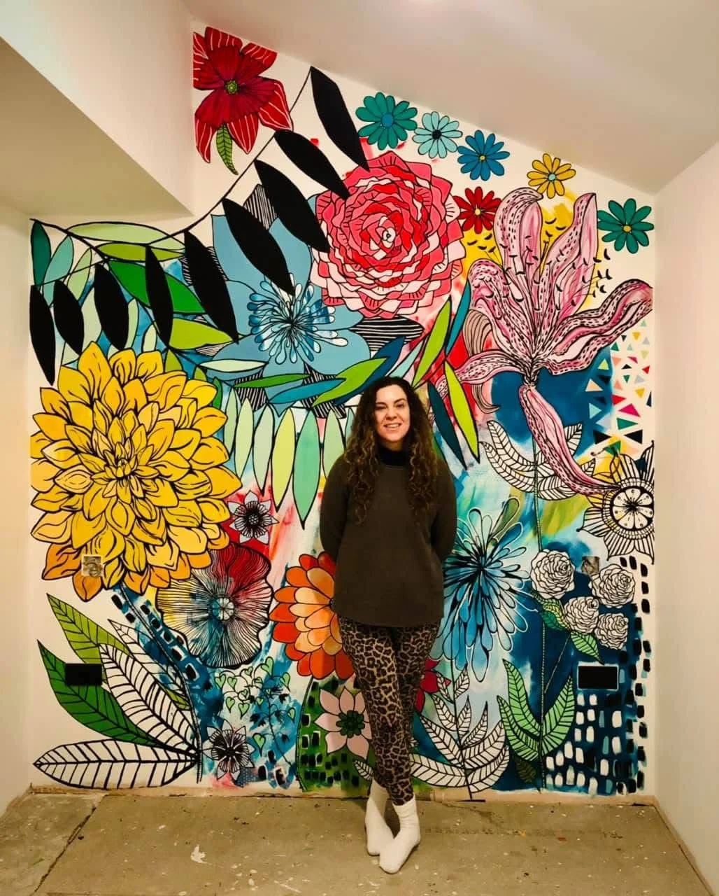 A woman with long curly hair wearing a black sweater, animal print pants, and white shoes standing in front of a vibrant painted wall filled with large, colorful flowers and leaves.