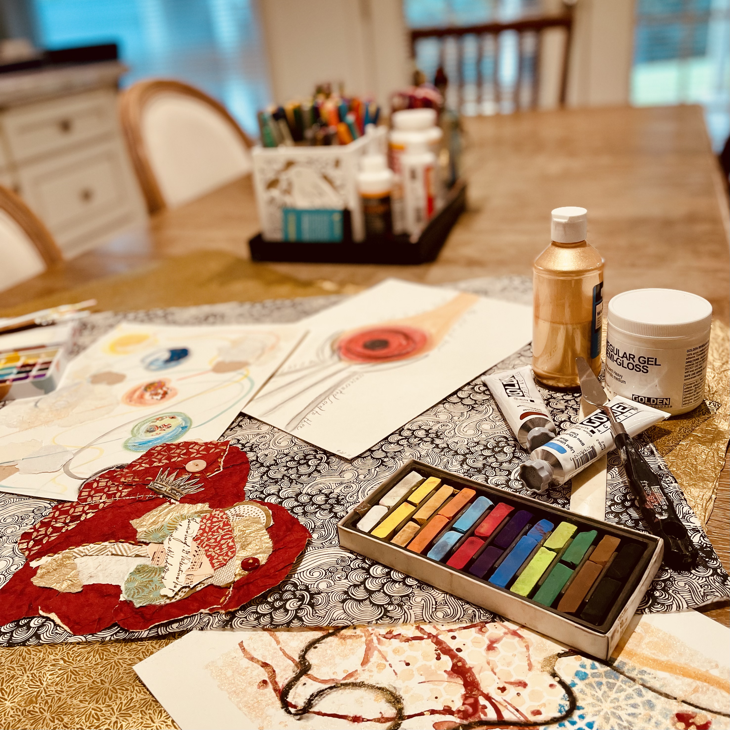 Art supplies on a table, including a box of colored pastels, watercolor paintings, bottles of gel medium, an orange bottle, and various paint tubes, with a wooden dining table and chairs in the background.