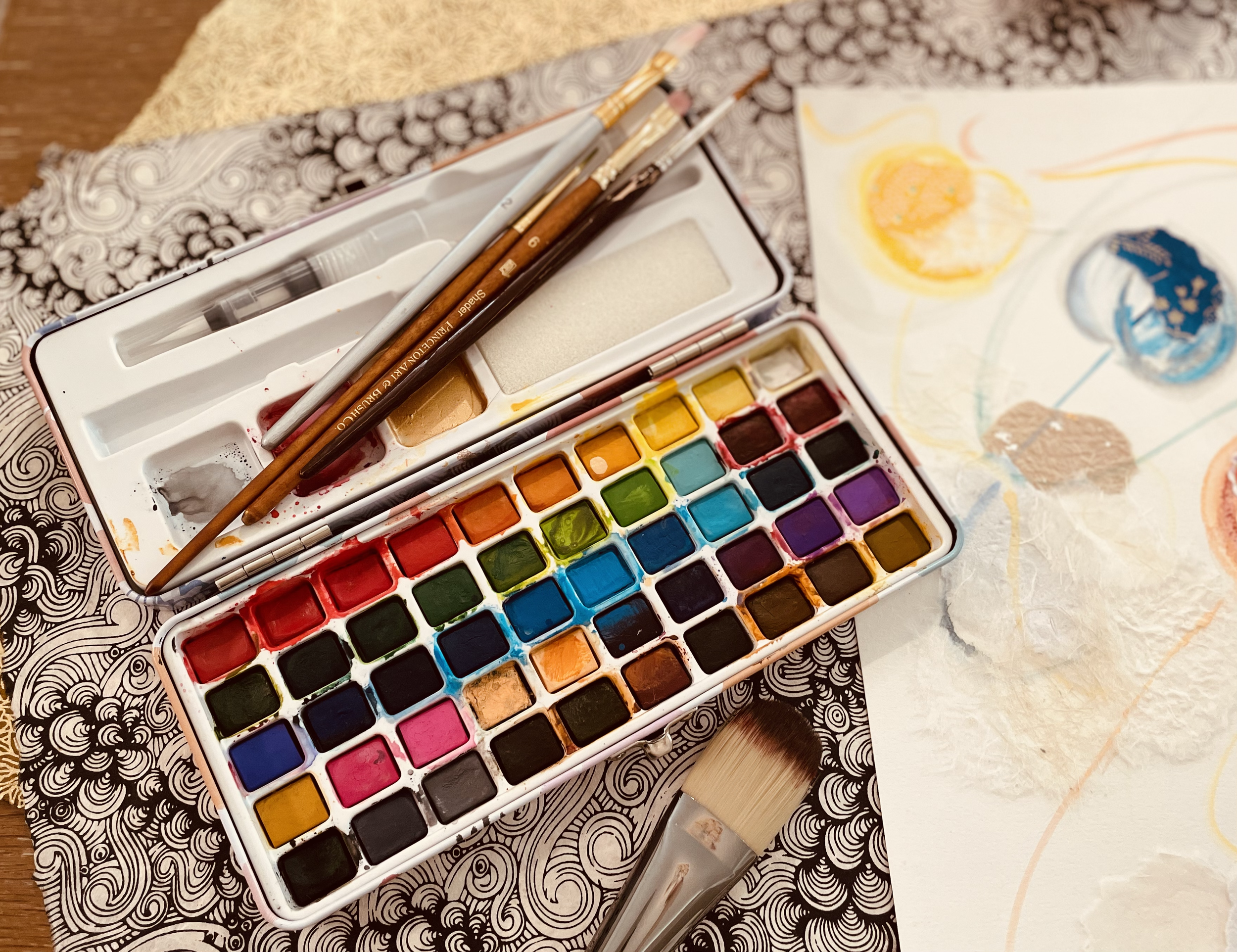 Open watercolor paint set with multiple colors, paintbrushes, and art supplies on a patterned surface. Some finished watercolor paintings of abstract circles are visible.