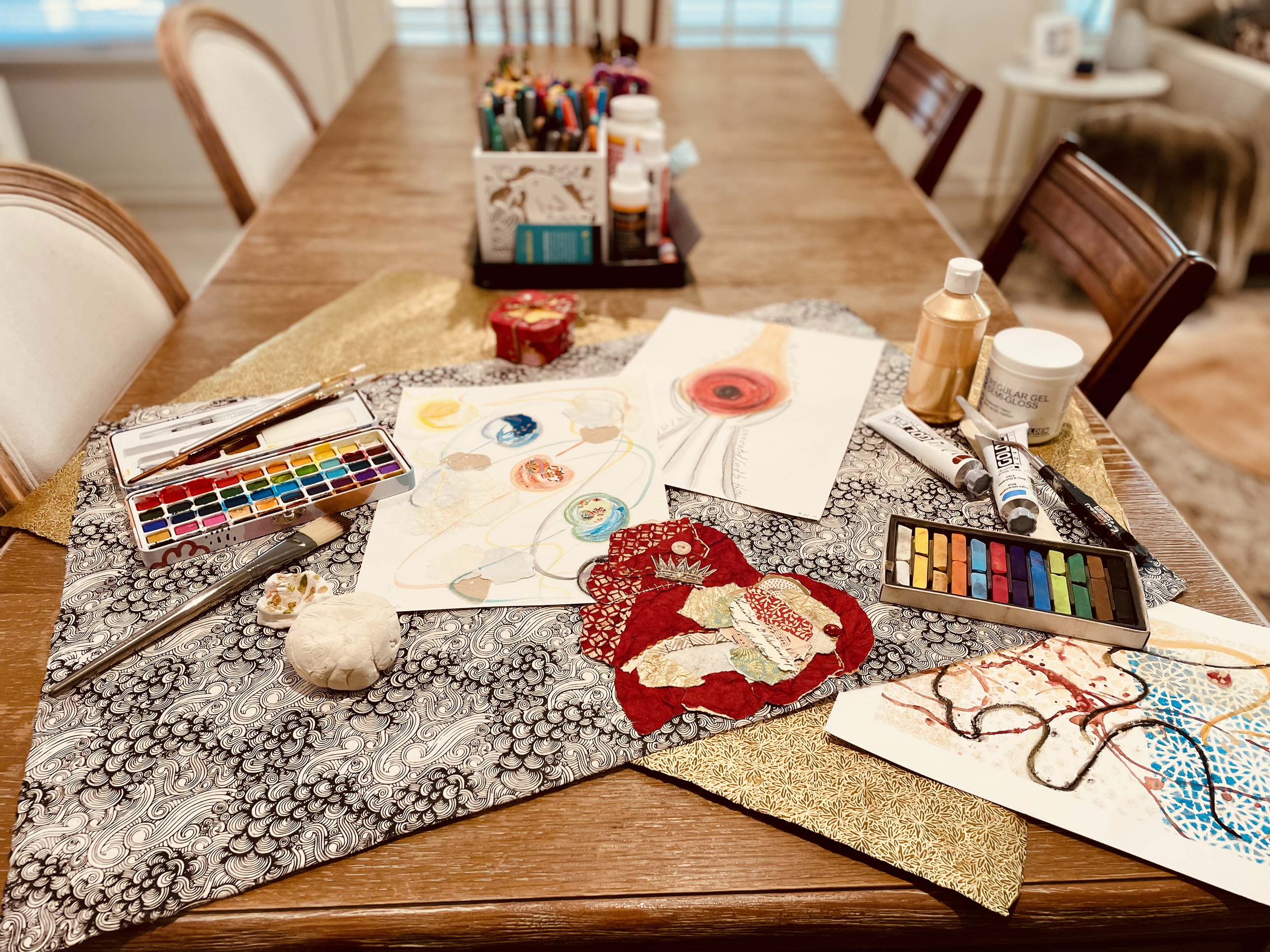 A wooden dining table covered with art supplies, including watercolor sets, brushes, and papers with colorful abstract paintings and fabric decorations, in a well-lit room with chairs.