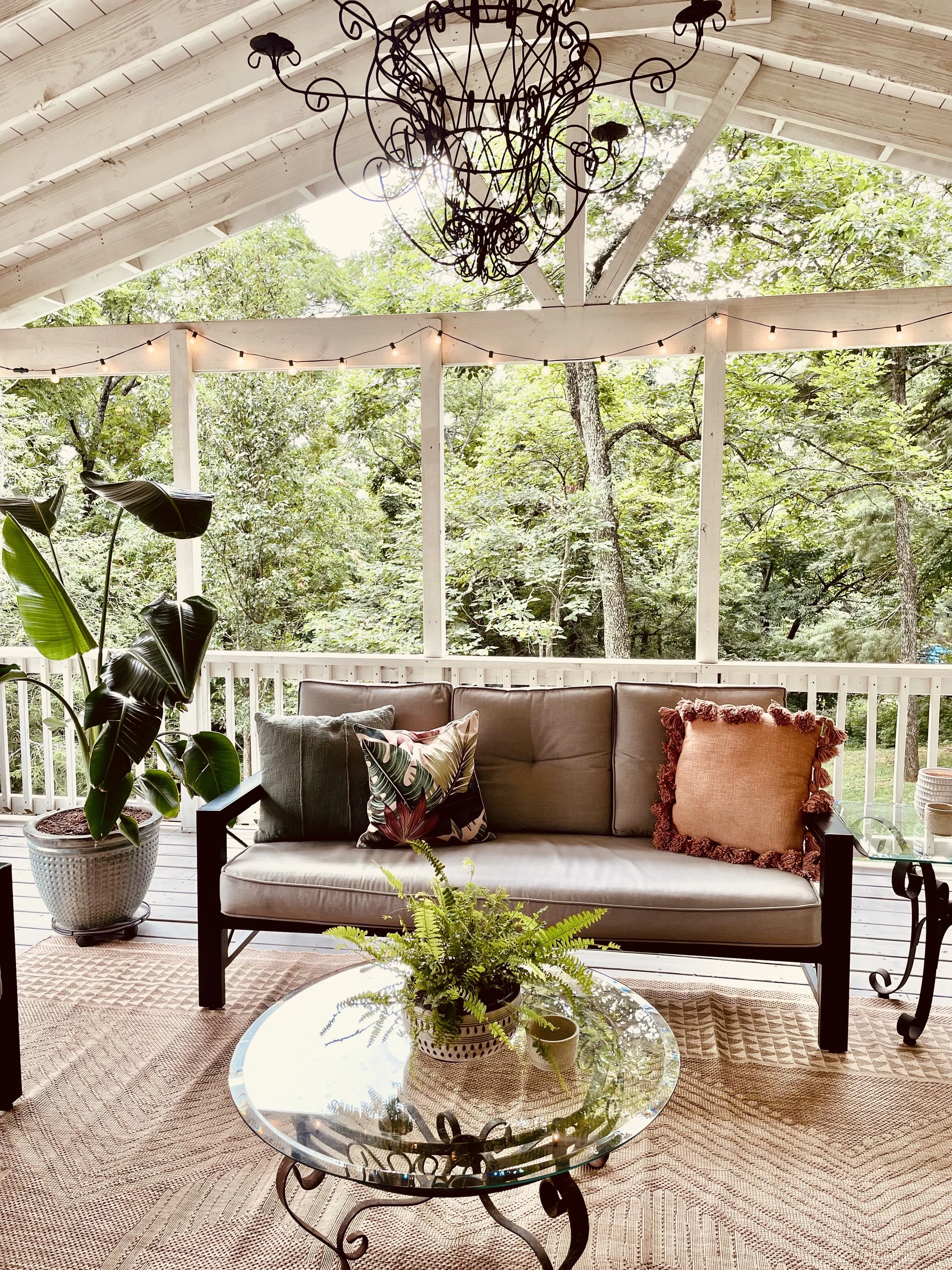 Covered porch with a gray cushioned sofa, colorful throw pillows, a potted plant on a glass-top coffee table, leafy plants, string lights, and a chandelier, with lush green trees in the background.