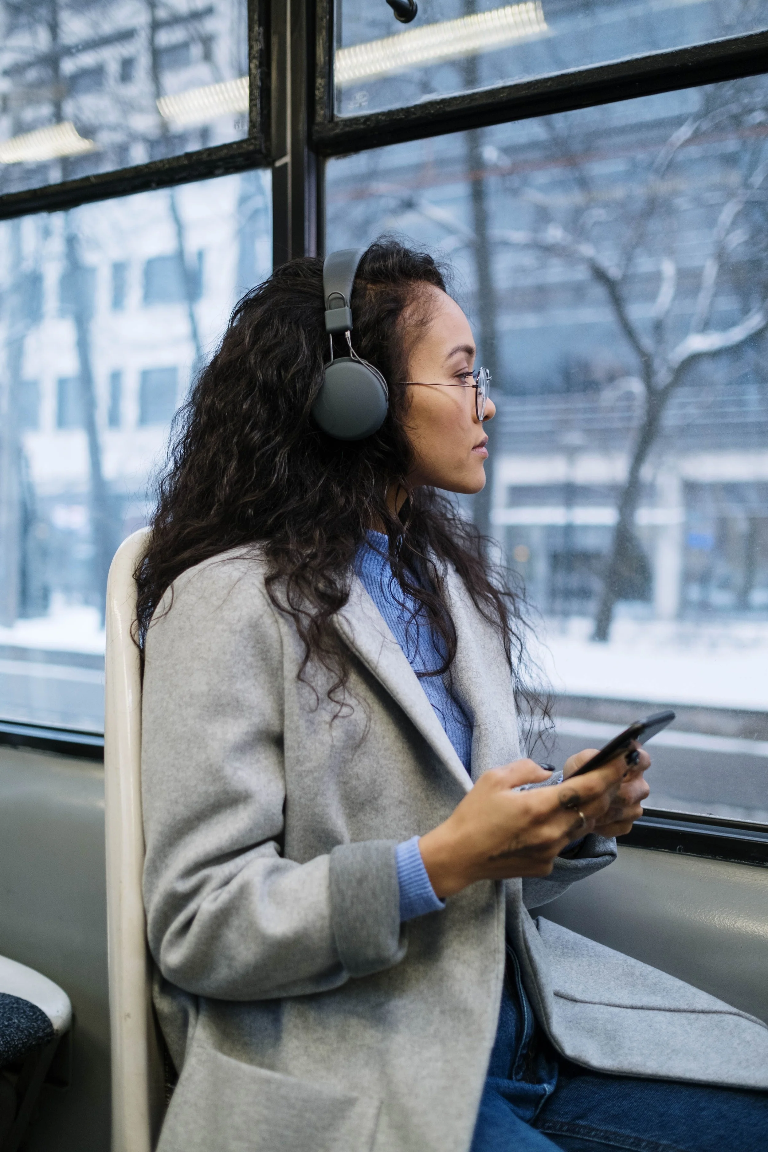 A young woman sits on public transit wearing headphones and looking at her phone, in a quiet moment in the middle of an ordinary day.