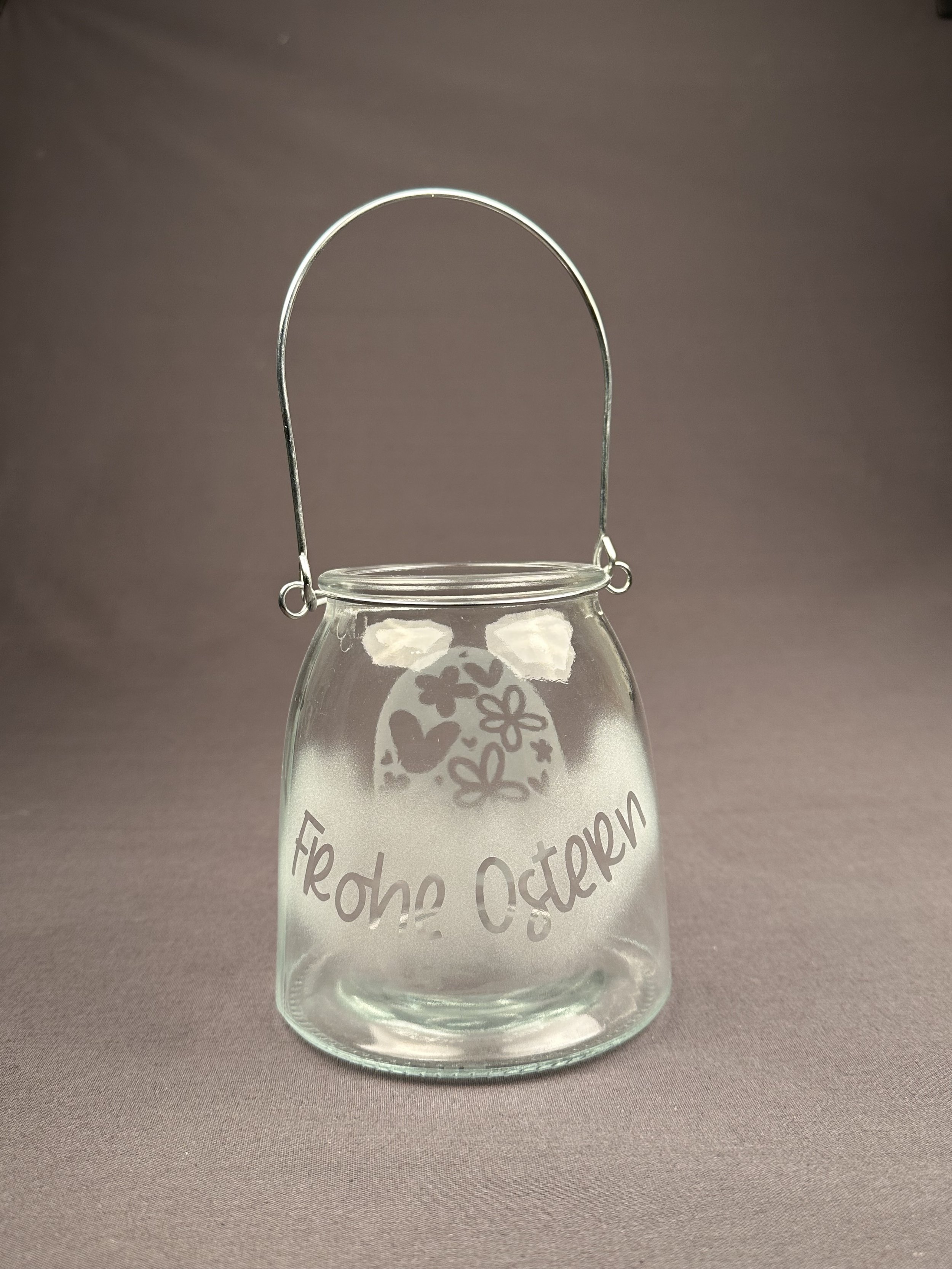 Ein kleiner Glaspilz mit einem Griff und der Aufschrift 'Frohe Ostern' im Deutschen. Es ist verziert mit einem Osterhasen, Ostereiern und Frühlingsblumen.