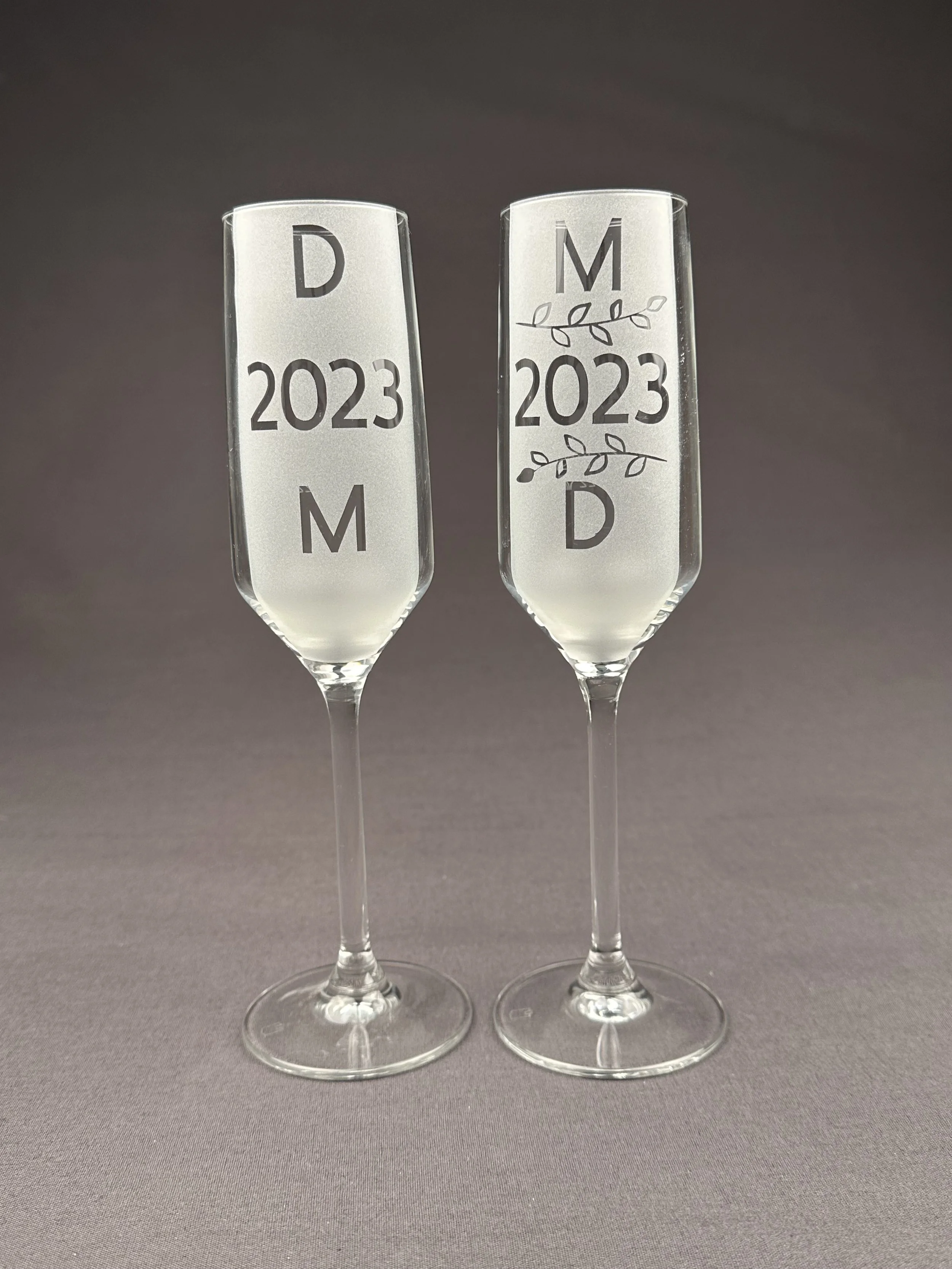 Zwei Champagnergläser mit gravierten Jahreszahlen 2023 und Buchstaben D und M, das rechte Glas mit einer dekorativen Blumen- und Blattgravur.