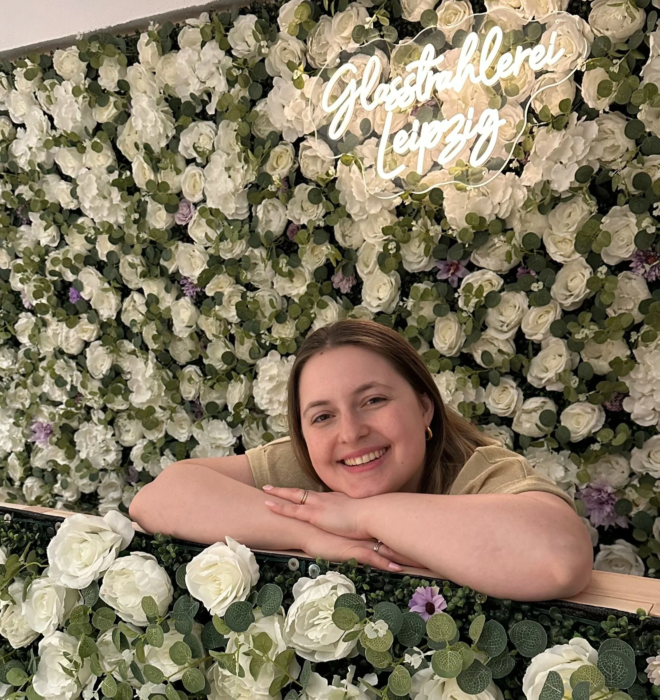 Junge Frau liegt mit den Armen auf einem Blumenpult aus weißen Rosen und grünen Blättern vor einer Wand aus weißen Blumen, im Hintergrund leuchtet ein Neon-Schild mit der Aufschrift "Glasstrahlerei Leipzig".