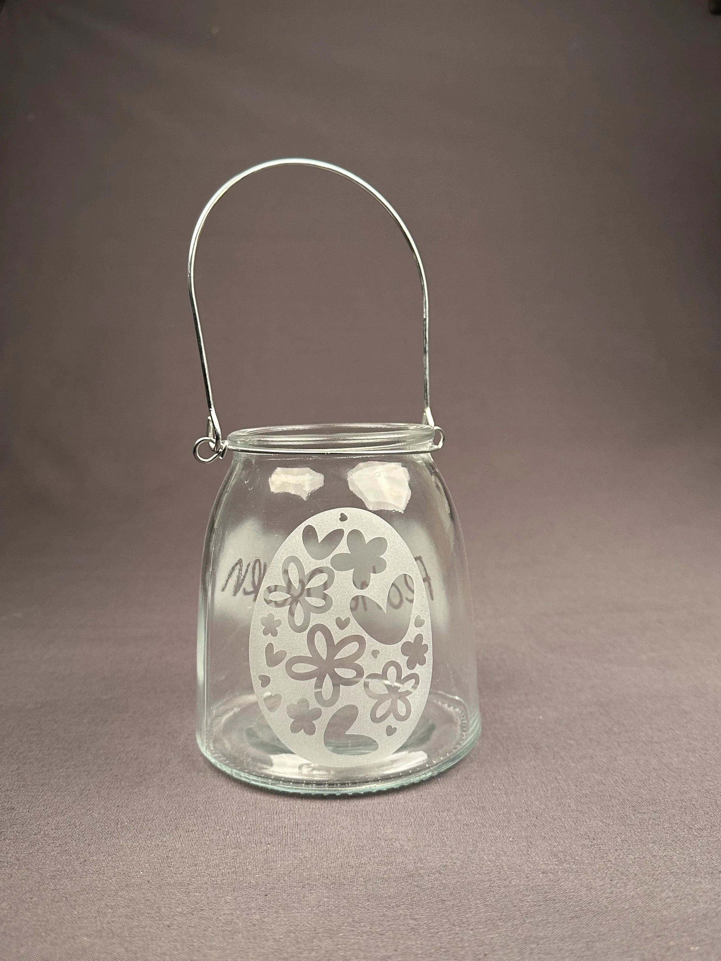 Ein transparentes Glas mit einem dekorativen Ei-Motiv aus Blüten und Herzen, mit einem Metallbügel zum Tragen.