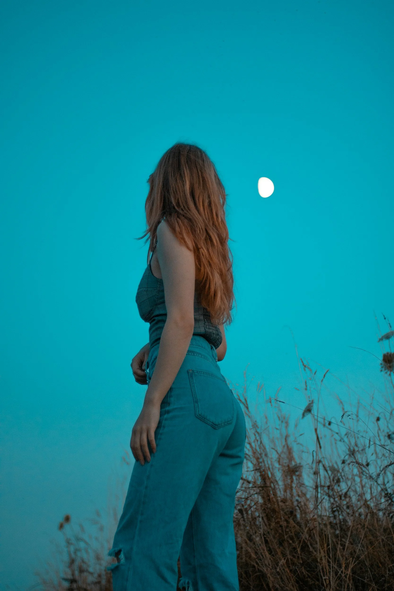 A woman with long red hair standing in a field at dusk, looking at the moon in the sky.