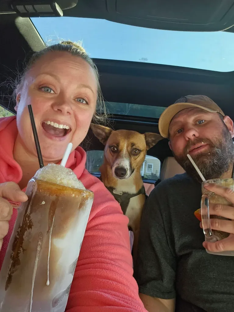 A woman, a man, and a dog inside a vehicle enjoying drinks, with the woman smiling widely showing her teeth, the man holding a drink, and the dog sitting between them looking at the camera.