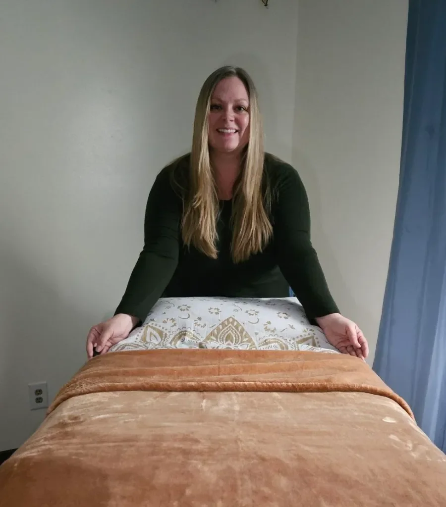 A woman with long blonde hair, wearing a black top, smiling and leaning over a massage table covered with a brown blanket.