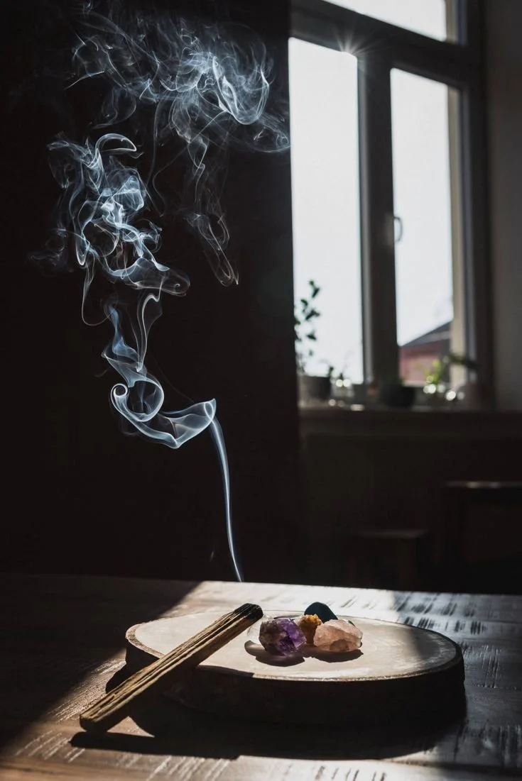 A wooden serving board with crystals and a pair of wooden chopsticks resting on it inside a room near a window, with smoke rising upwards.