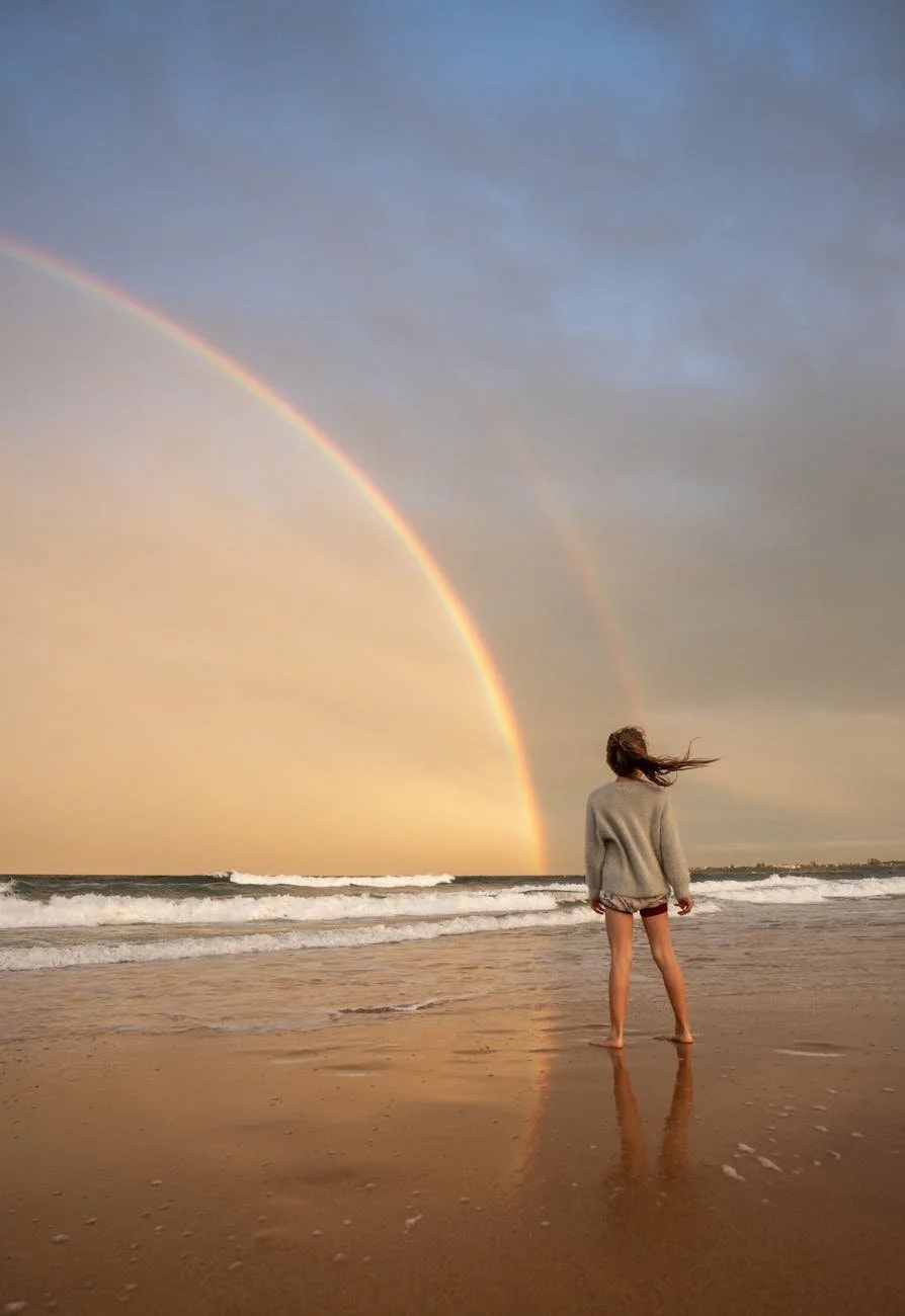 A woman standing on a beach facing the ocean with a rainbow and a double rainbow in the sky, wind blowing her hair and a cloudy sky at sunset.