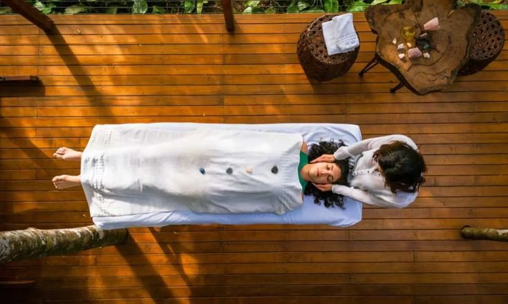 A person lying on a massage table covered with a white sheet, receiving a Reiki healing treatment from a practitioner. The scene is set on a wooden deck with outdoor furniture and greenery in the background.