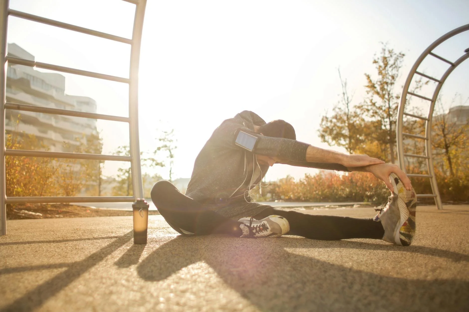 Person doing a seated hamstring stretch outside during sunrise, wearing athletic clothes and sneakers, with a water bottle nearby.