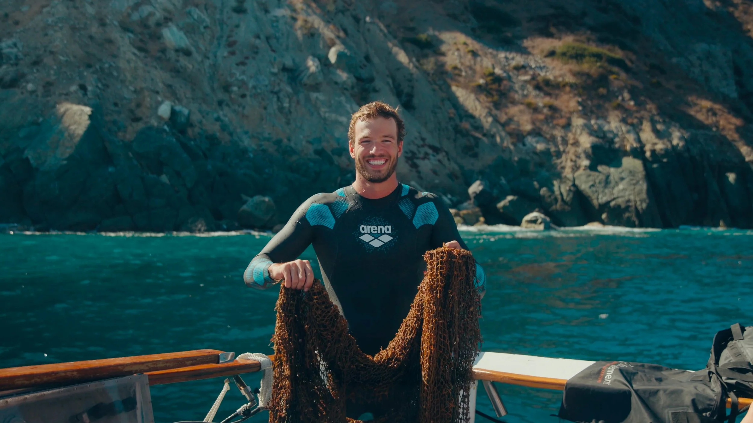 A man in a wetsuit on a boat holding a brown fishing net, smiling with a rocky coastline and ocean in the background.