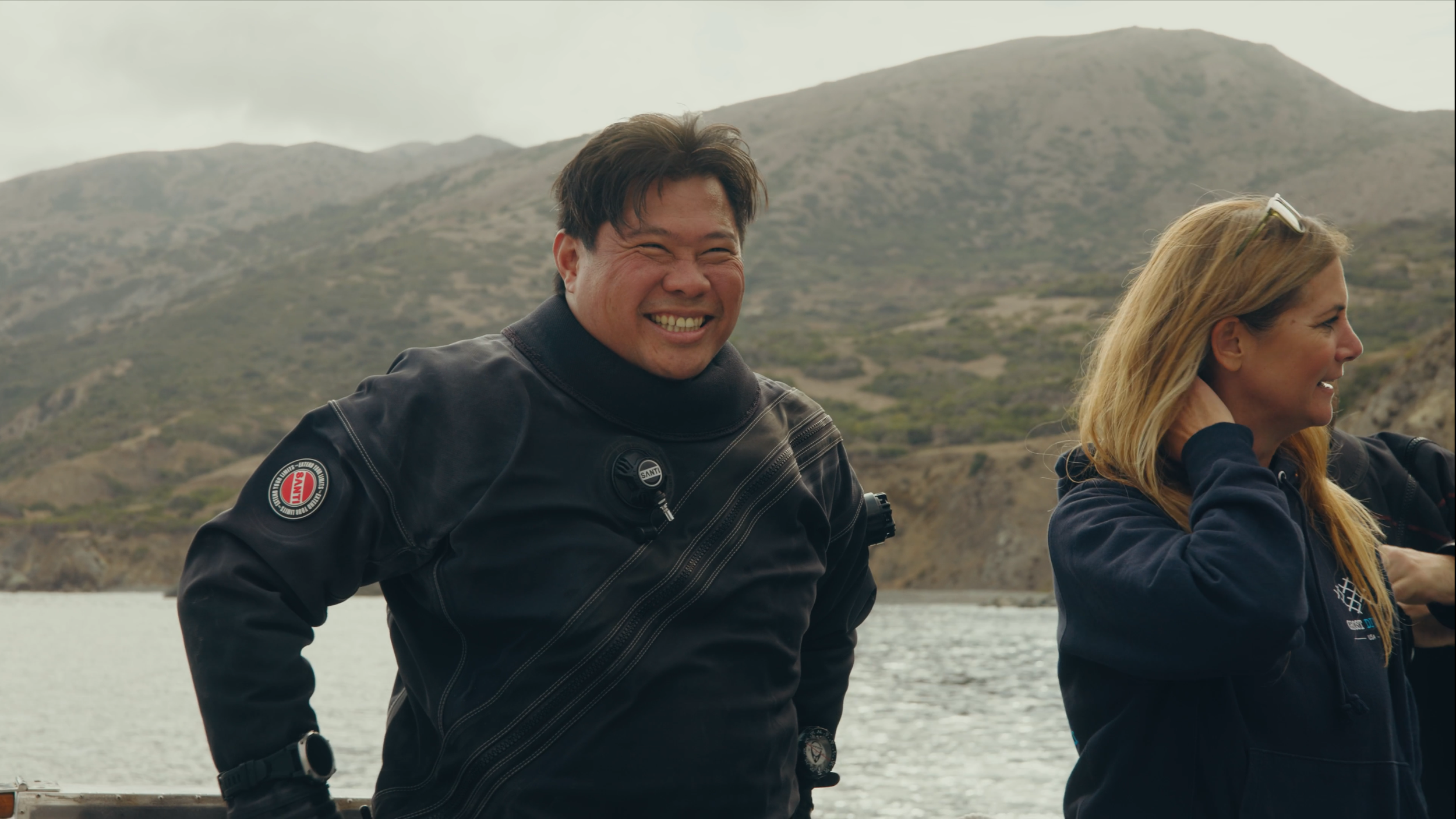 Two people outdoors near a body of water with mountains in the background, smiling and dressed in jackets suitable for cool weather.