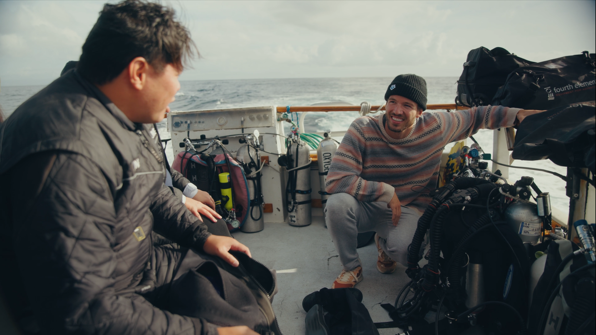 Two men are on a boat, one seated and one squatting, surrounded by scuba gear and dive tanks, with the ocean in the background, engaging in conversation.