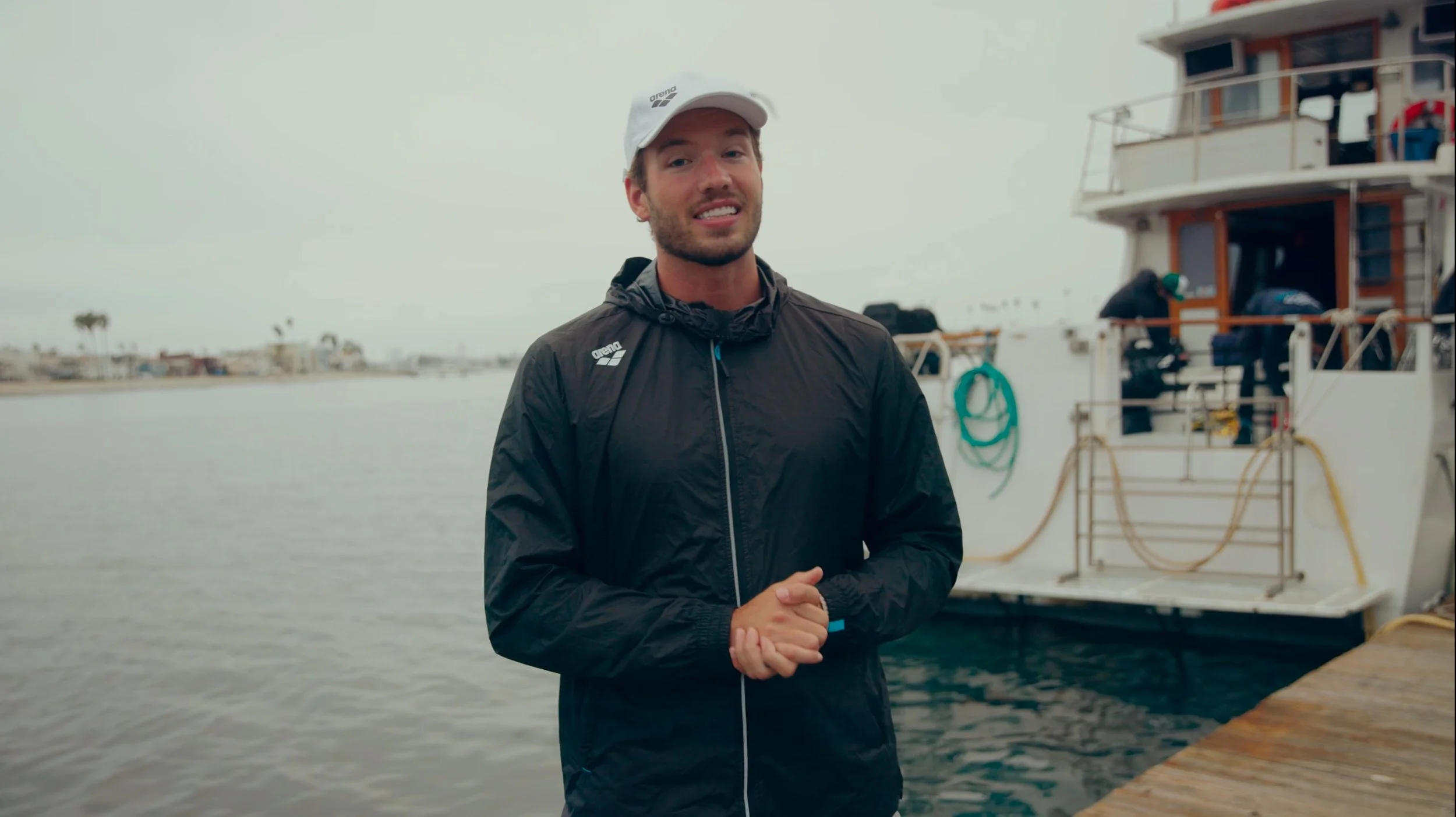 A man with a beard and smiling, wearing a white cap and black jacket, stands near a dock with a boat in the background on a cloudy day.
