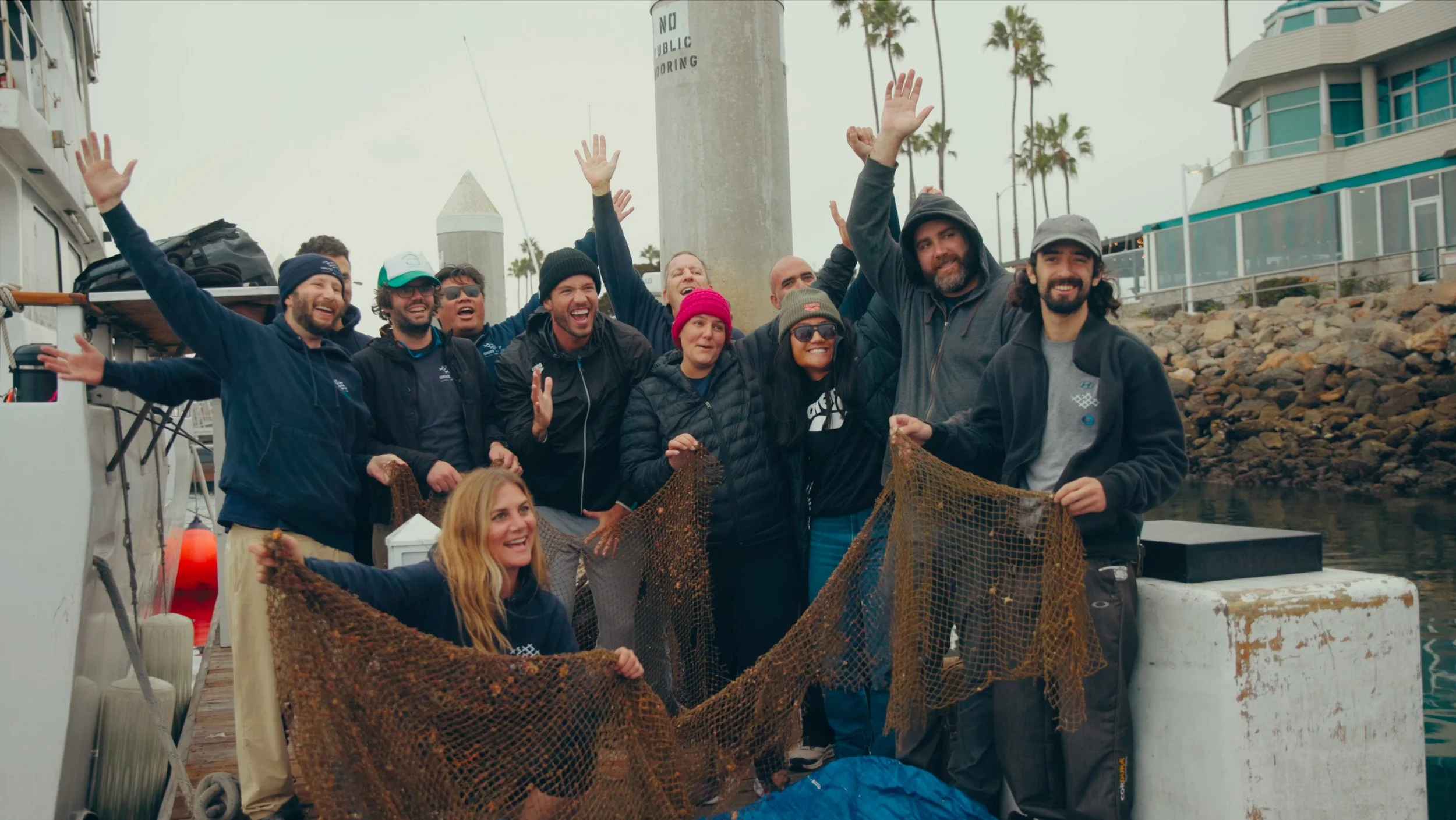Group of people on a dock celebrating with a fishing net, with a boat nearby and palm trees in the background.