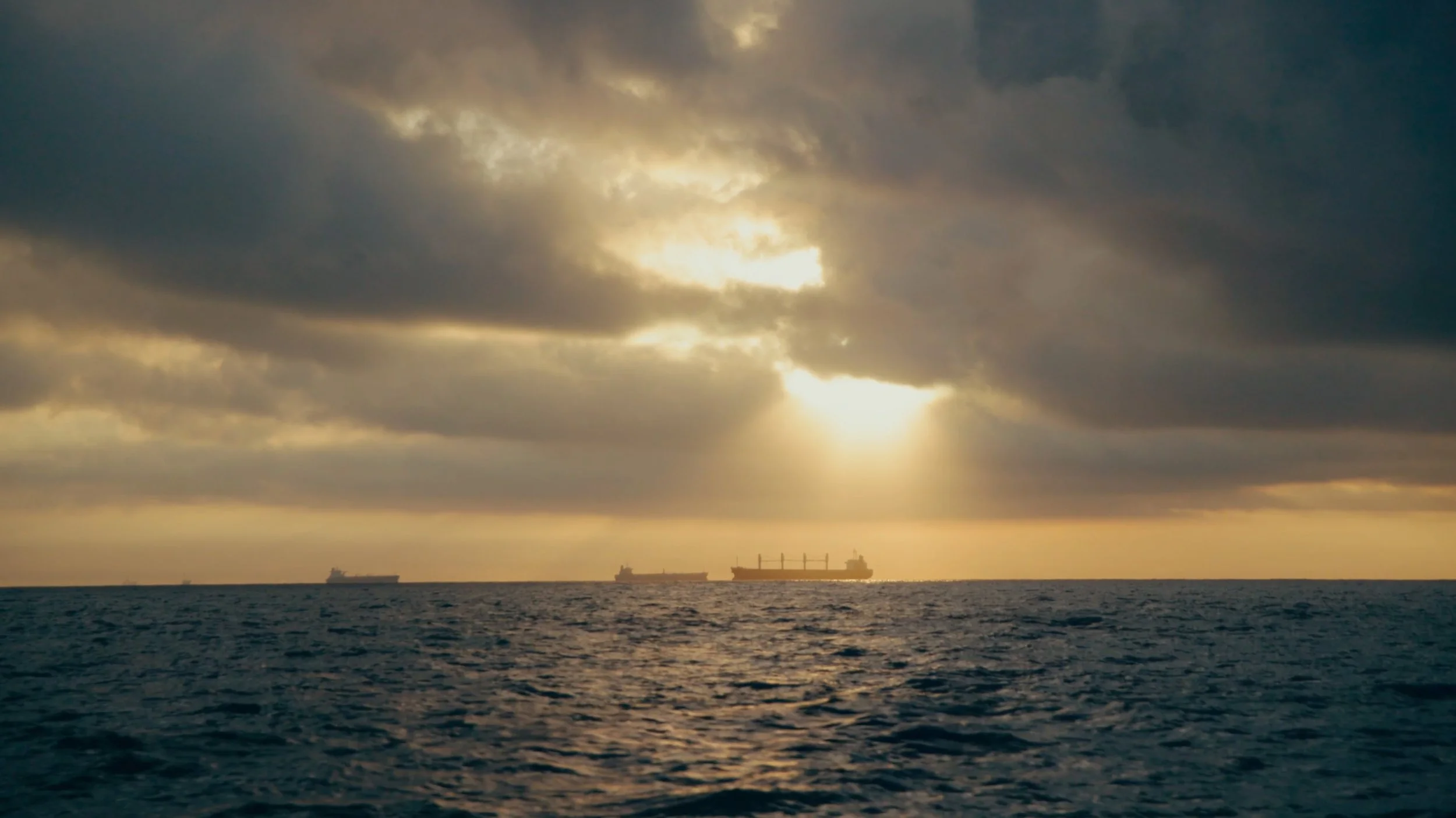 Sunset over the ocean with several cargo ships in the distance, partially obscured by clouds.